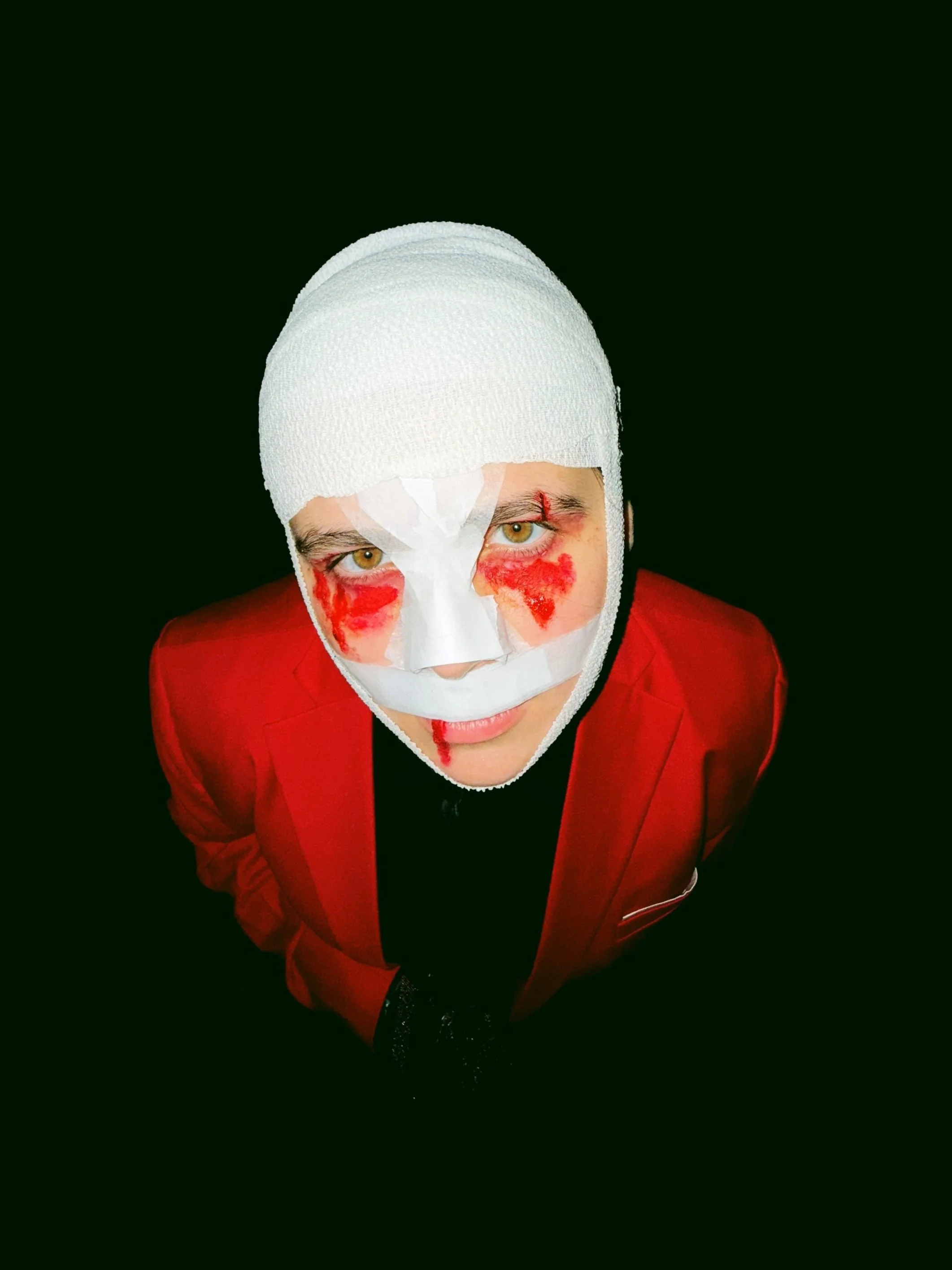 Person with face painted in red and white, wearing a white bandage headgear and a red blazer, looking directly at the camera against a black background.