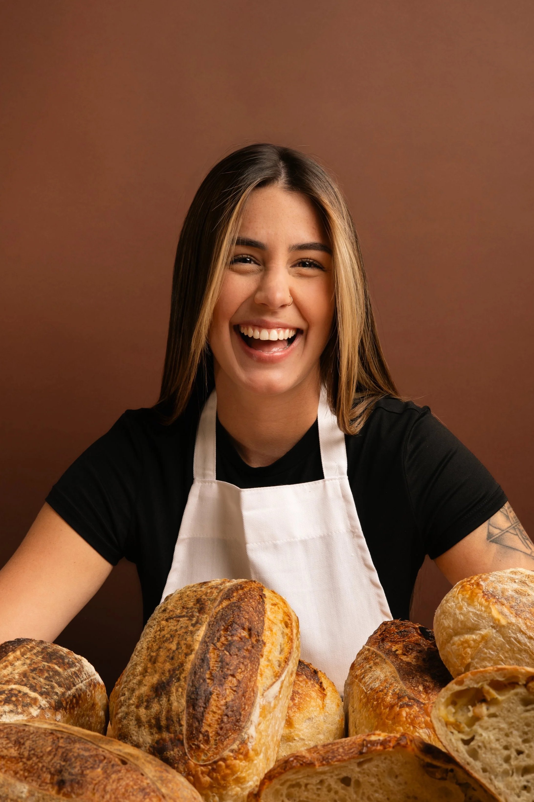 A smiling woman with long dark hair, wearing a black shirt and white apron, surrounded by various loaves of bread against a brown background.