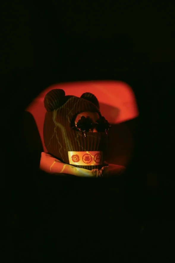 A stuffed animal wearing sunglasses and a band with symbols, illuminated by a red light in a dark environment.