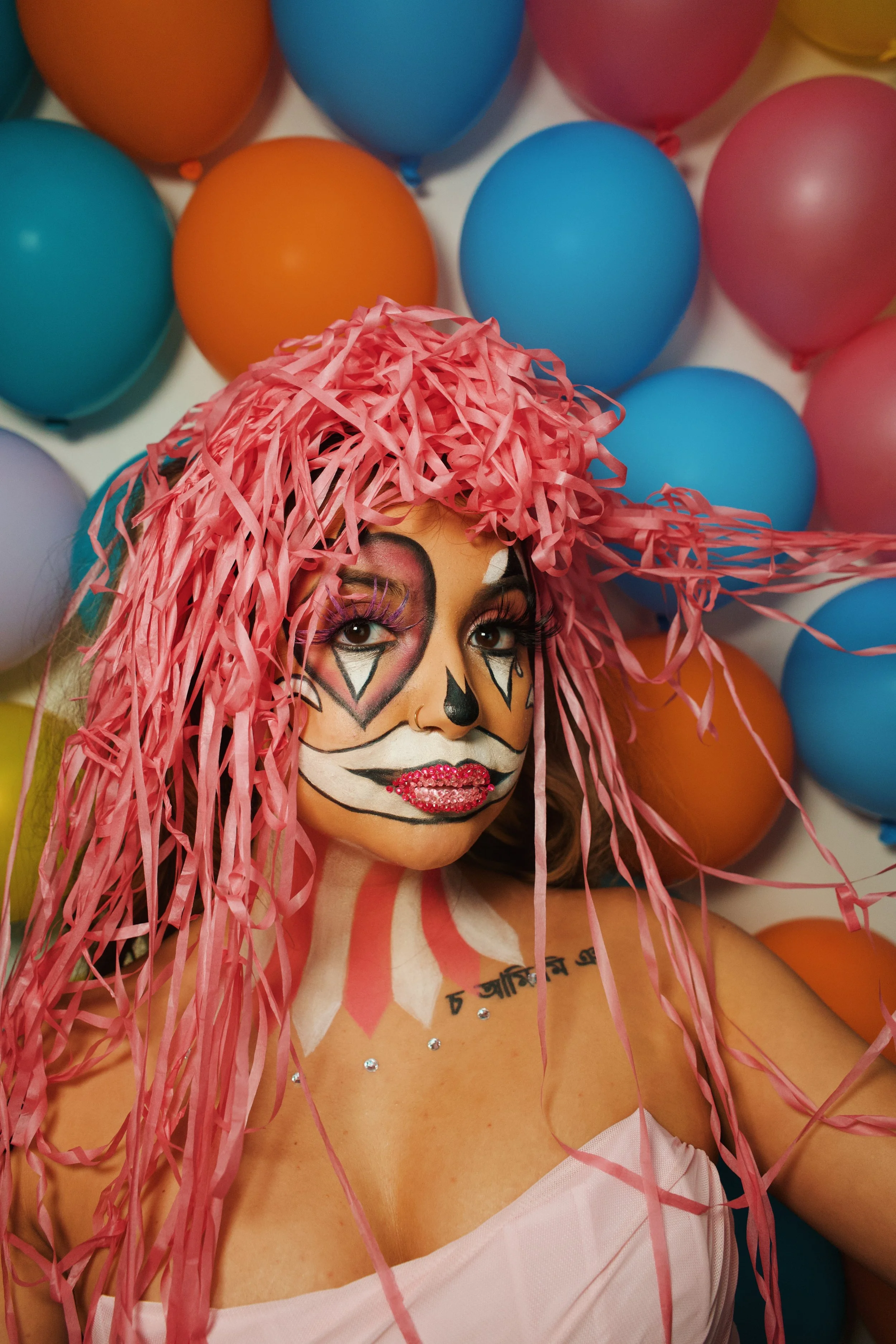 Person wearing colorful sugar skull makeup and pink shredded wig, surrounded by multicolored balloons.