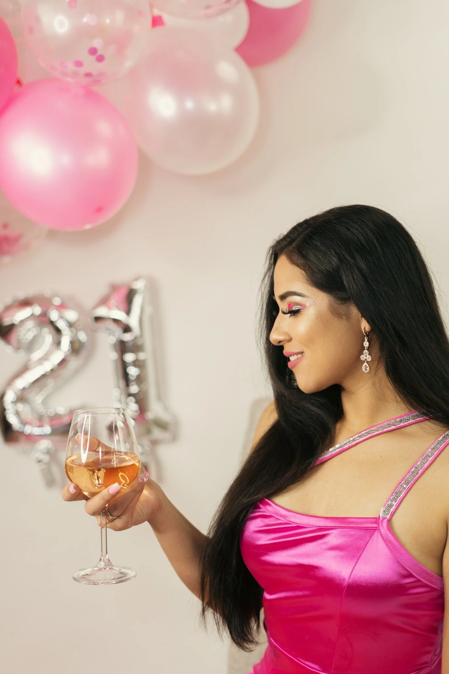 A woman in a pink dress holding a glass of wine during a celebration party with pink balloons and numbered silver balloons in the background