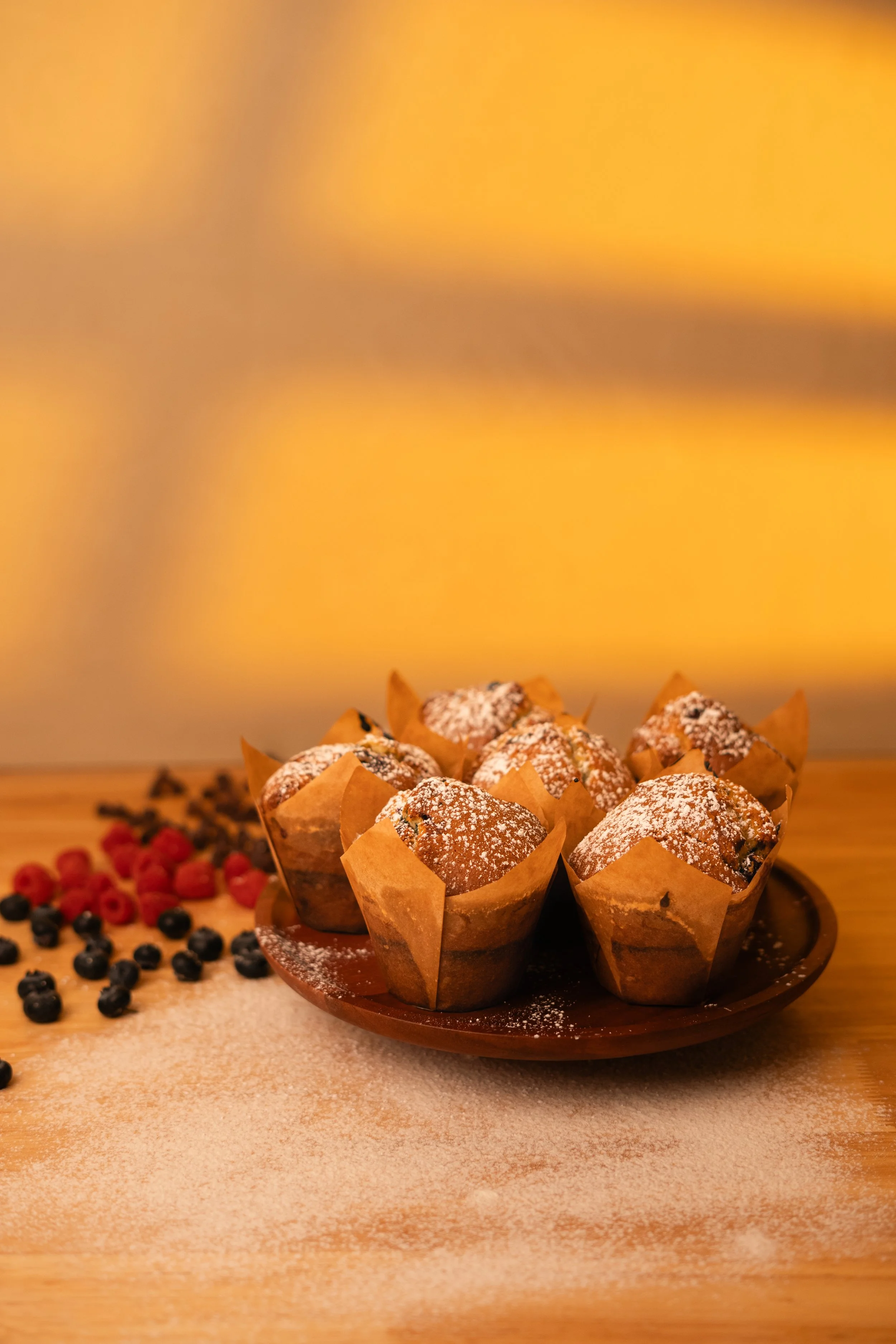 A plate of muffins sprinkled with powdered sugar, with an assortment of berries scattered around on a wooden surface, against a yellow background.