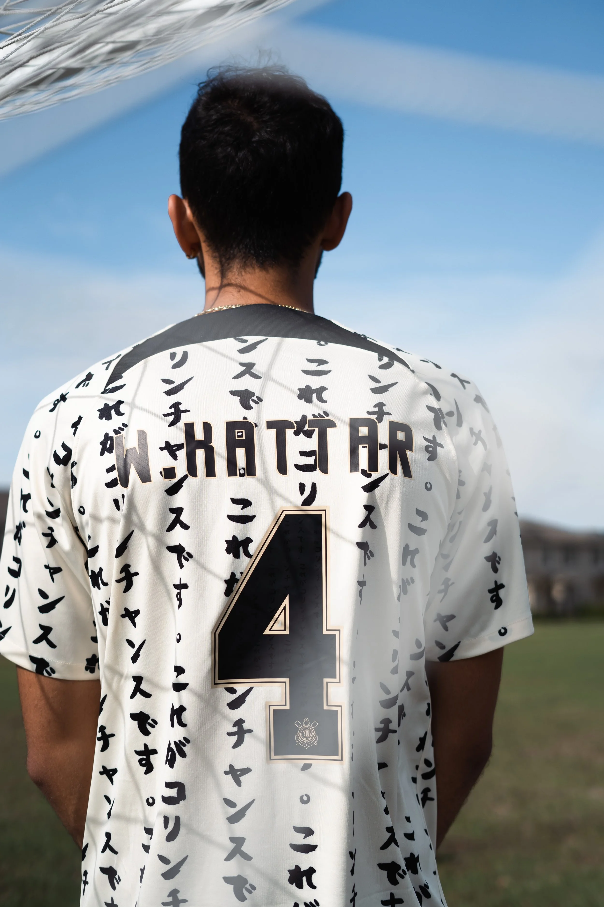 Person with short dark hair viewed from the back, wearing a white sports jersey with black lettering and the number 4, standing outdoors on grass with a blue sky and some buildings in the background.