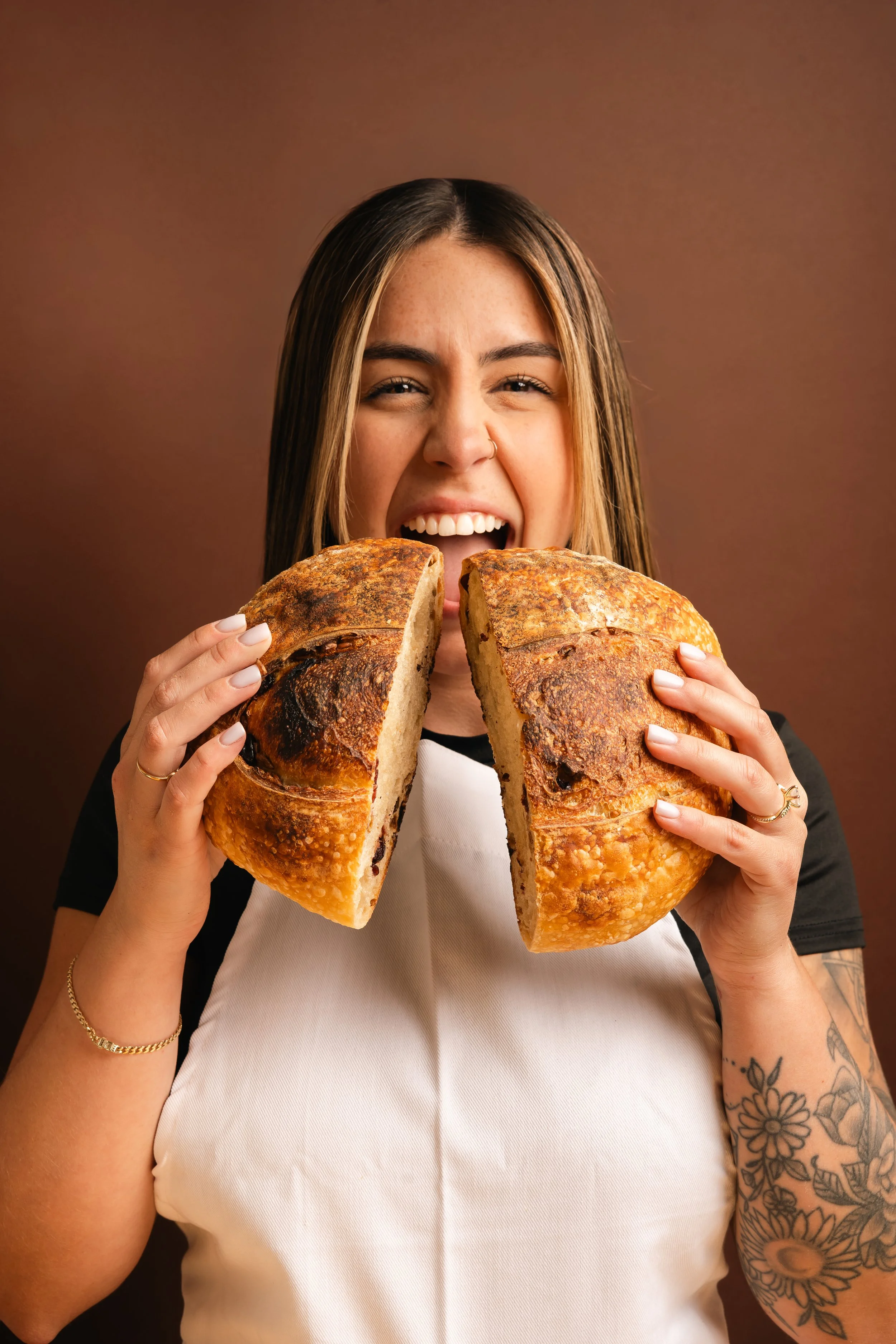 A woman with long hair, tattoos on her arm, and jewelry is holding a large, partially sliced loaf of bread and smiling.