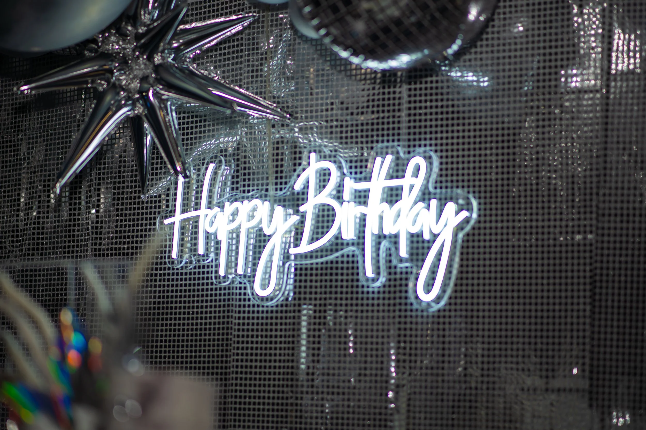A neon sign that reads 'Happy Birthday' in bright white cursive letters, with metallic star-shaped balloons and decorations around it.
