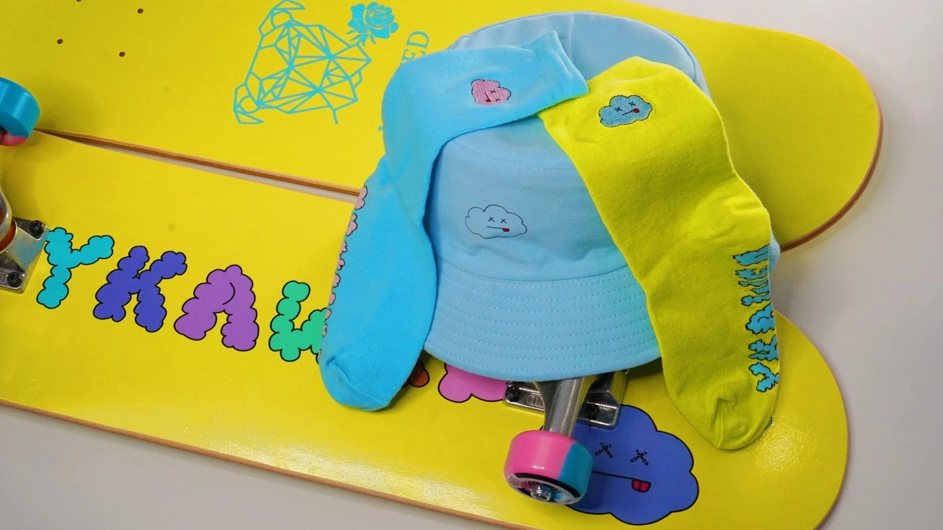 Colorful skateboard with yellow deck, decorated with cloud and bubble graphics, topped with blue and yellow socks and a blue cap, all featuring small embroidered cloud patches, resting on a white surface.