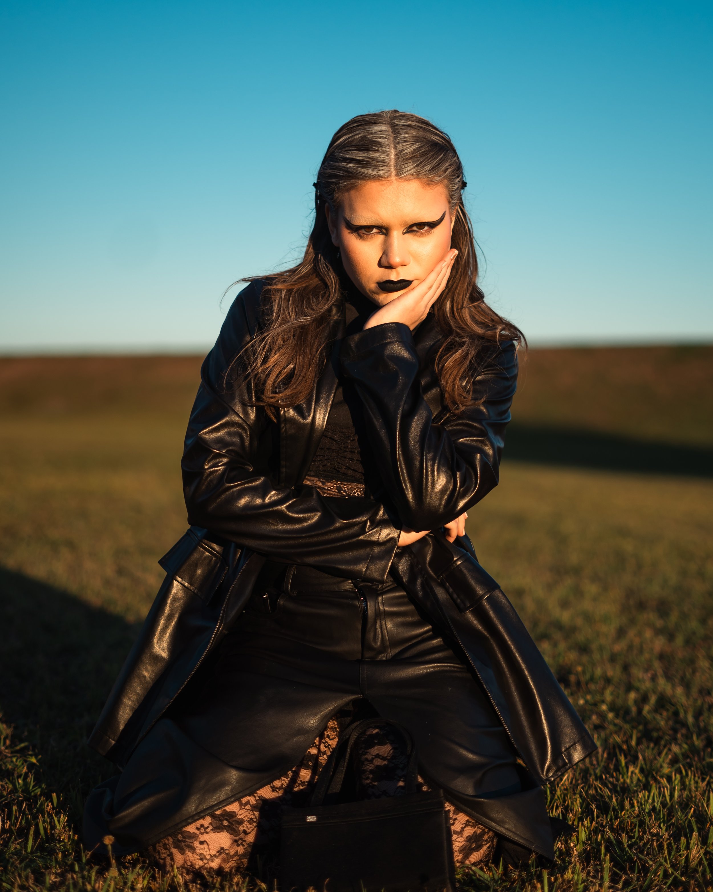 A young woman with dramatic makeup, dressed in black leather, kneeling on grass in an open field during sunset.