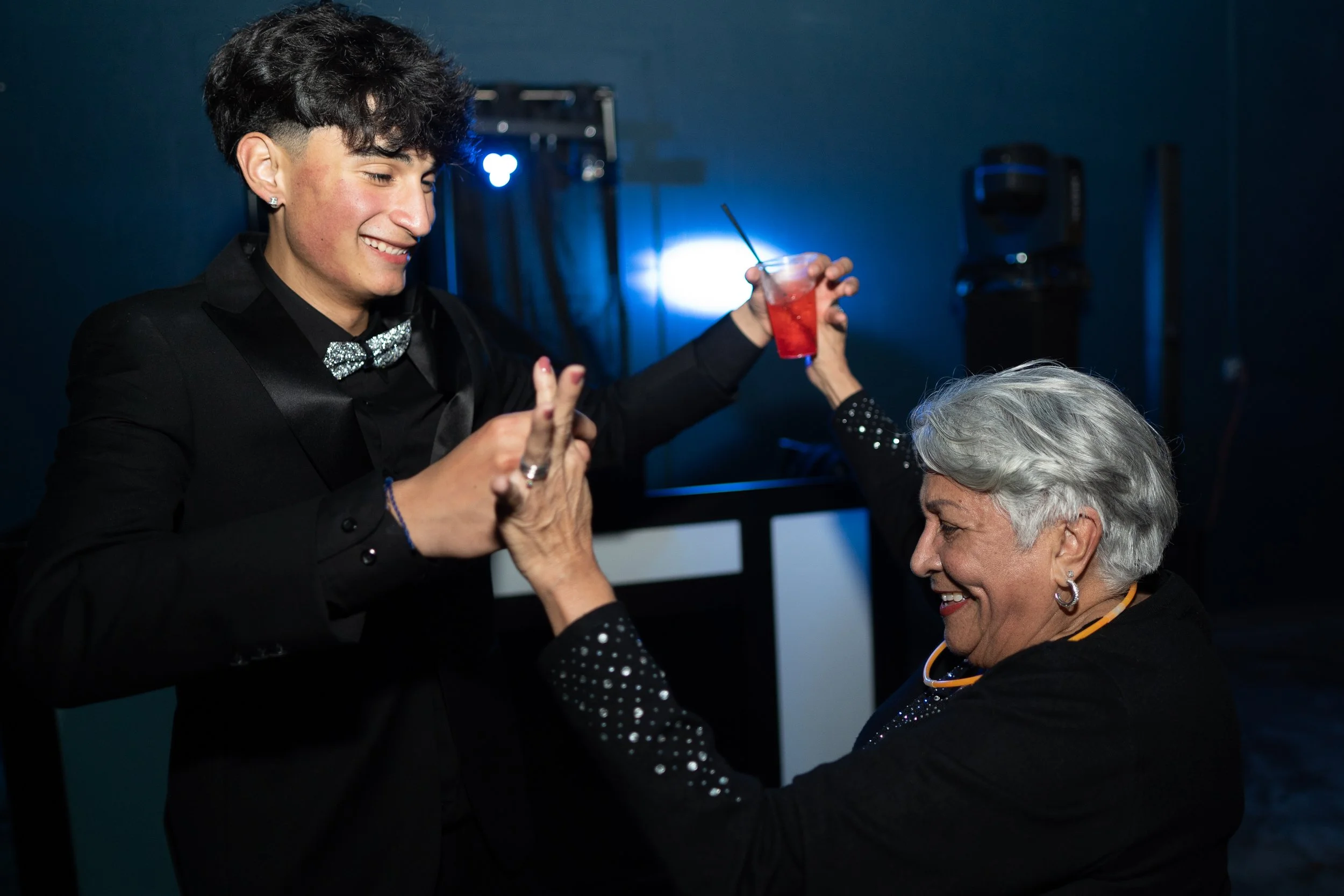 A young man in a black suit and silver bowtie dancing and smiling with an older woman with white hair, who is also smiling and holding a red drink, in a dark room with blue lighting.