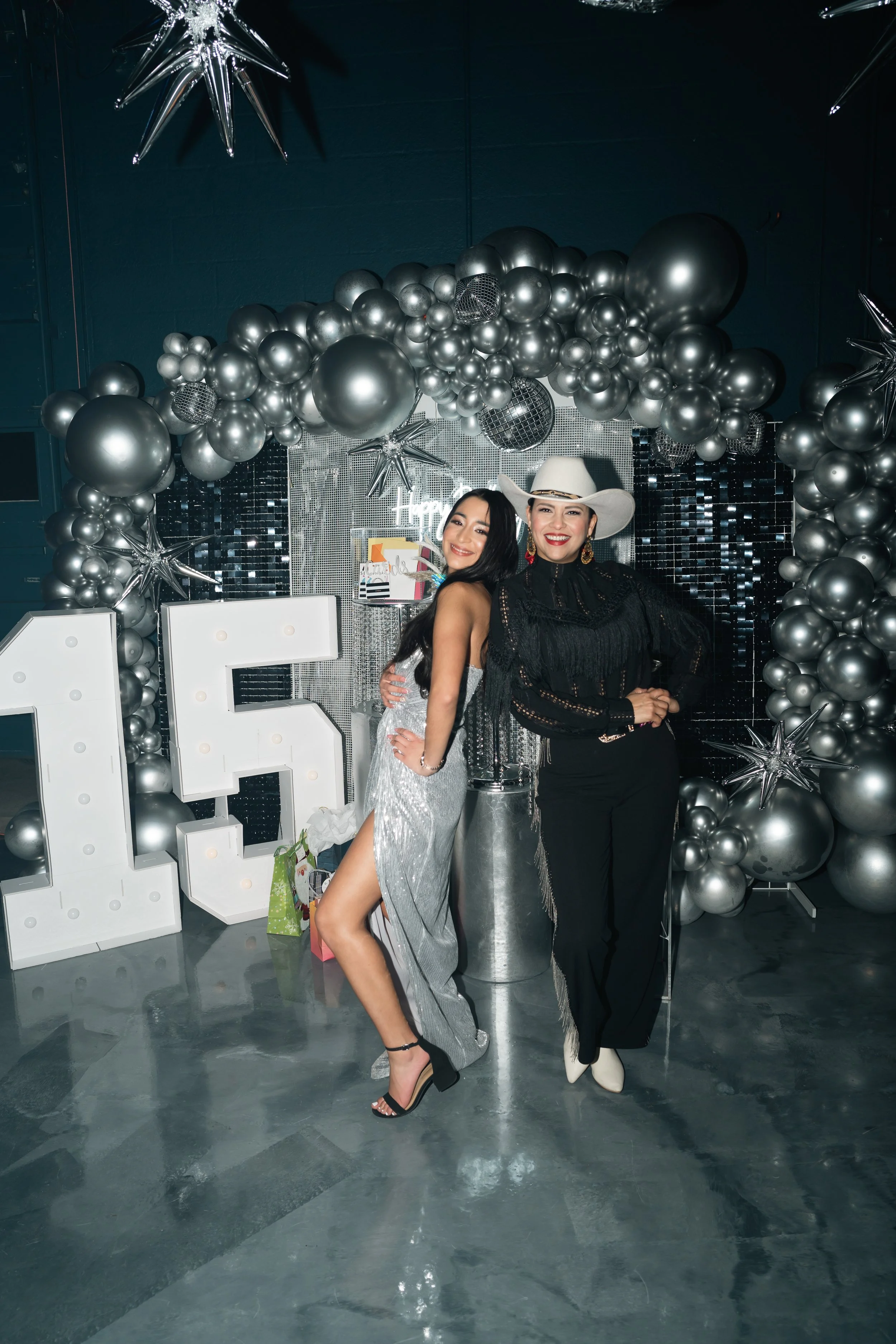 Two women posing at a celebration with silver balloons, large illuminated number 15, and silver star balloons, indicating a 15th birthday or anniversary celebration.