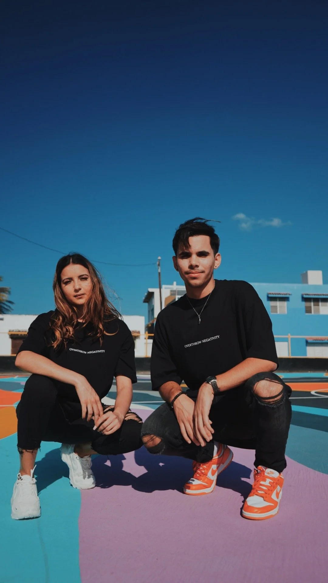 Two young adults, a woman and a man, squatting on a colorful outdoor mural in front of a blue building under a clear blue sky. They are wearing black t-shirts with white text and casual black pants, with the man wearing orange and white sneakers.