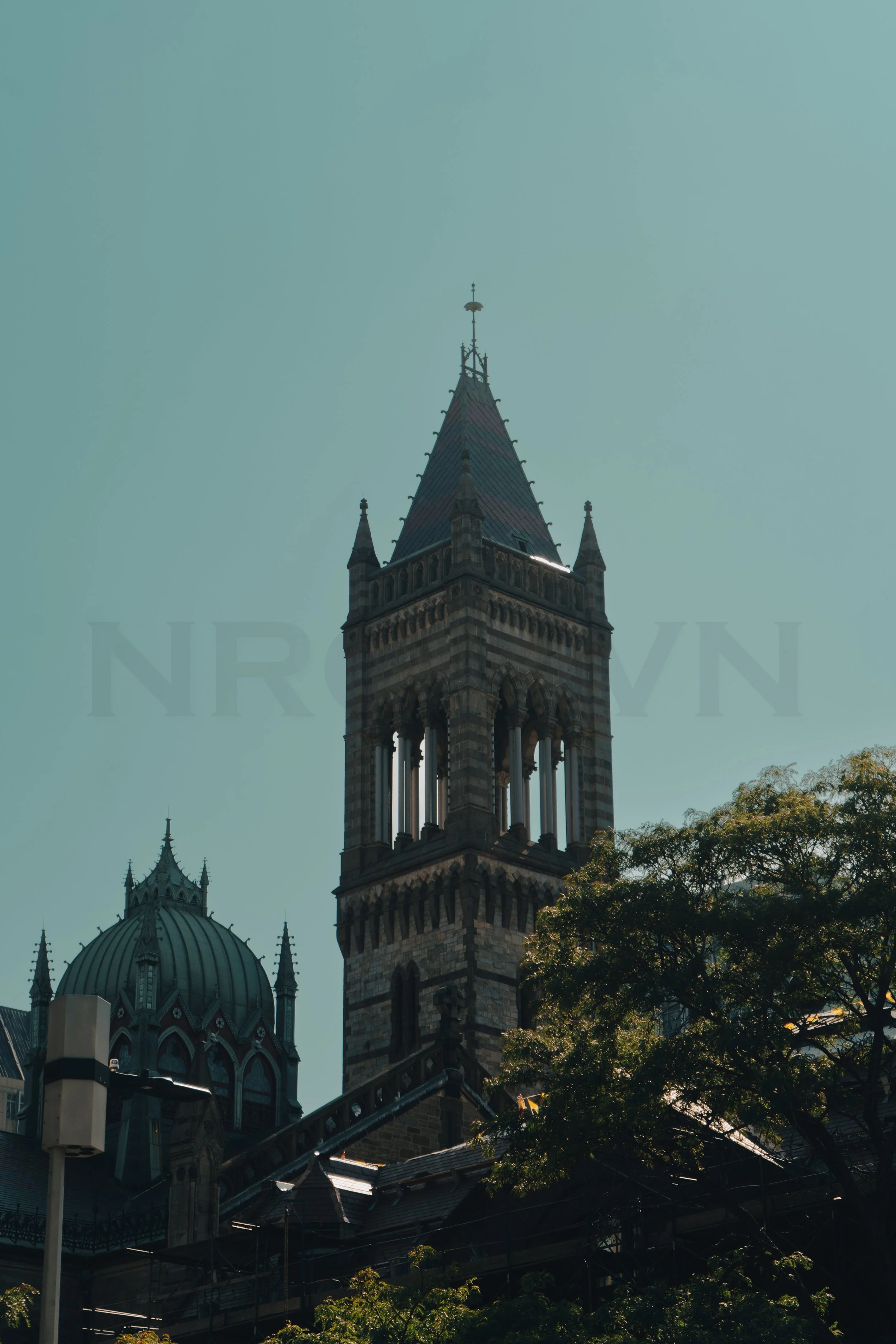 A tall, historic clock tower with pointed spire and detailed architecture, partially obscured by green trees, with a clear sky in the background.