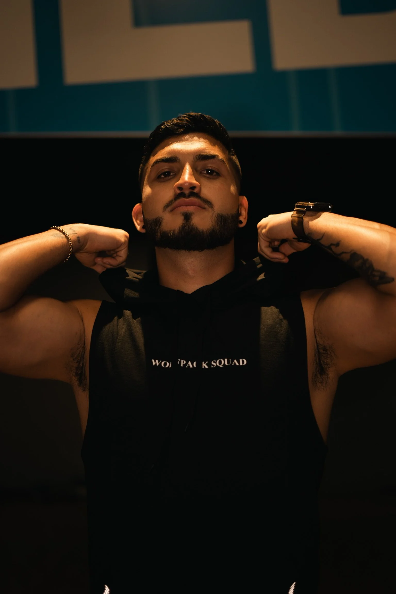 A man with a beard and tattoos adjusting his sleeveless hoodie in a dimly lit room.
