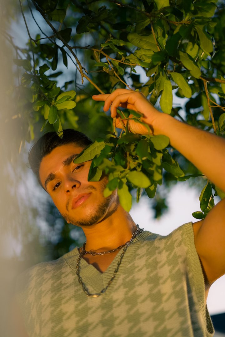 Young man with dark hair and beard in a sleeveless patterned shirt and layered necklaces, standing outdoors, among green leafy branches, during the evening.
