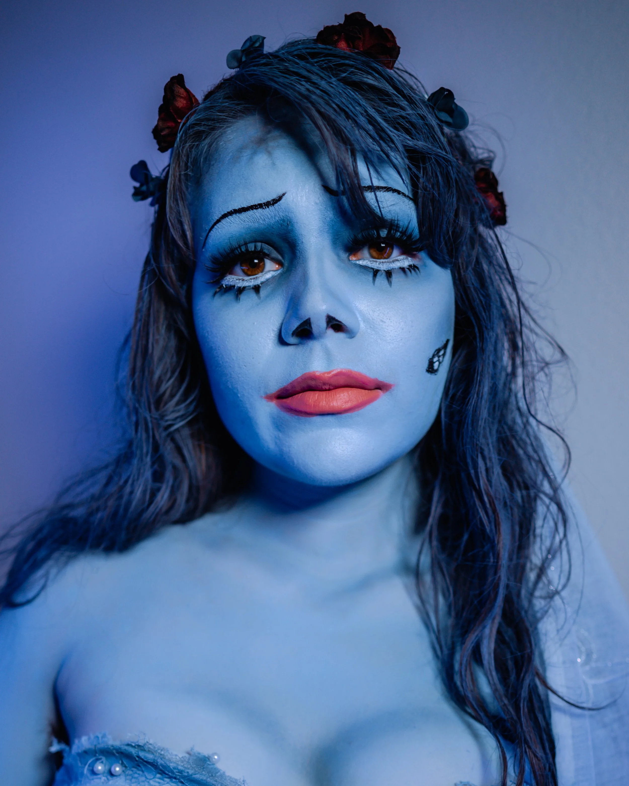 A woman with blue skin and artistic makeup resembling a skeleton face, with flowers in her hair, looking directly at the camera.