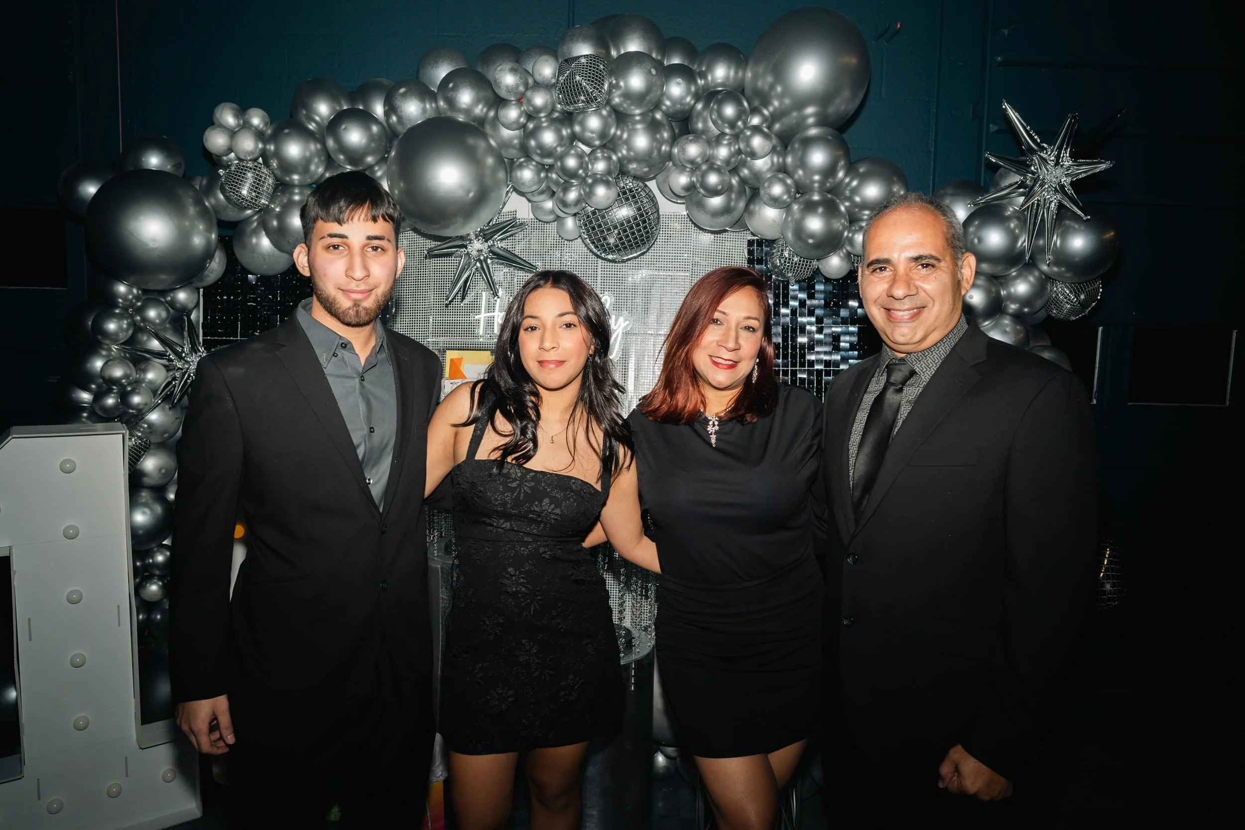 Four people dressed in formal attire posing in front of a silver balloon arch at a celebration event.
