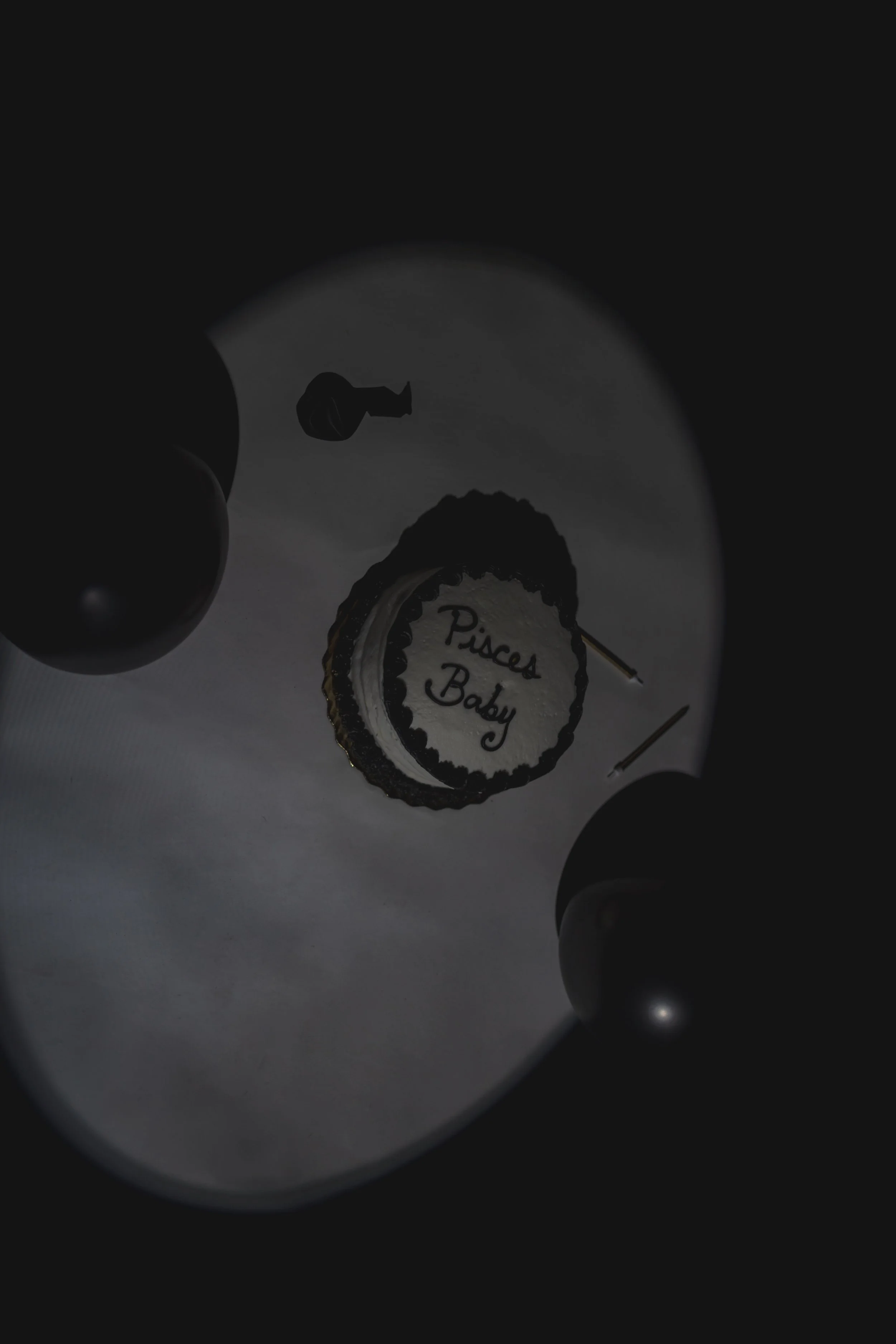 A birthday cake with the words 'Pisces Baby' written on it, flanked by two candles, viewed through a dark reflective surface, with balloons partially visible.