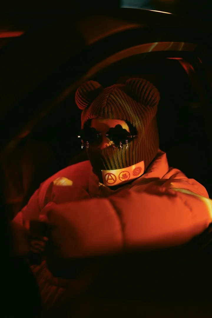 Person wearing a beanie with bear ears, sunglasses, a face mask, and a puffy jacket, illuminated by red and orange lighting.