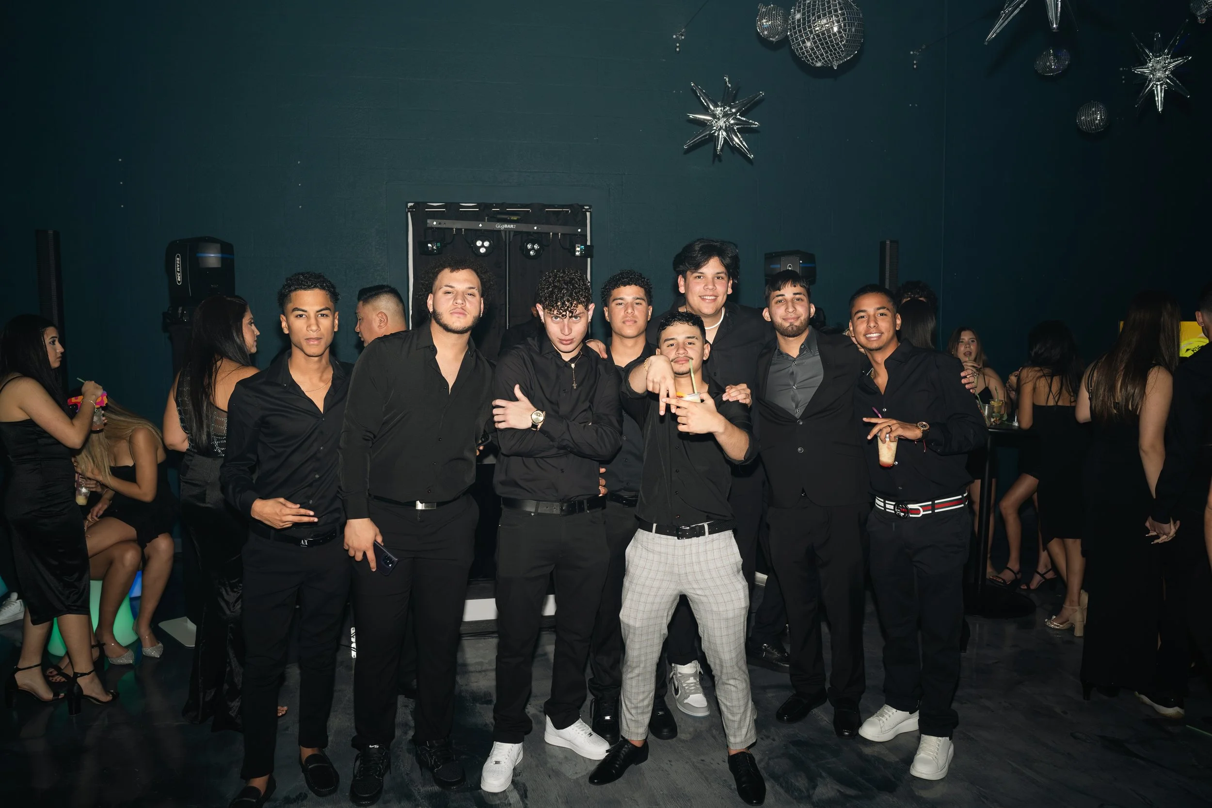 Group of young men dressed in black at a party, posing for a photo in a dimly lit room with silver star-shaped and disco ball decorations hanging from the ceiling.