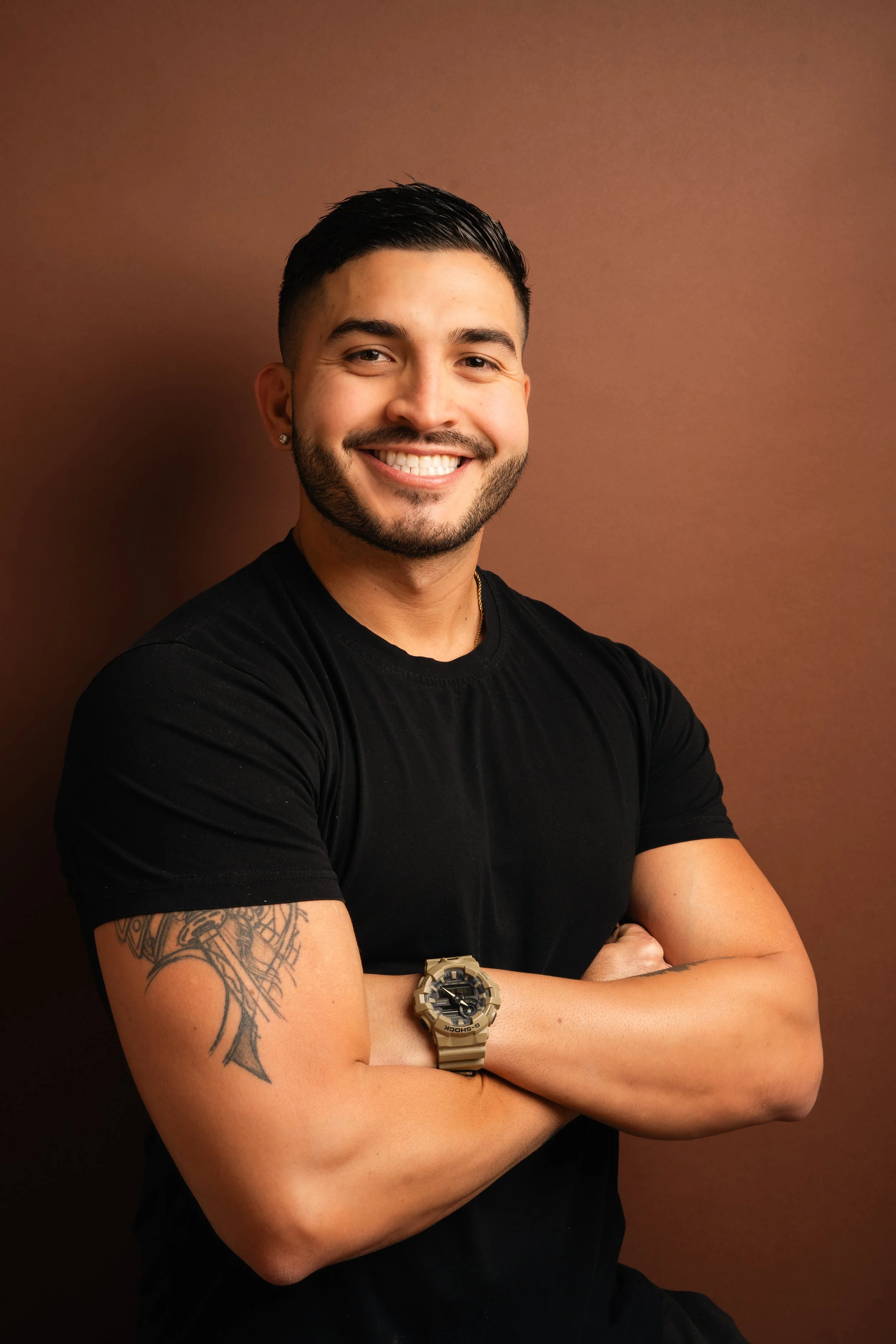 A smiling man with dark hair, a beard, earrings, and a tattoo on his arm, wearing a black t-shirt and a watch, standing against a brown background.