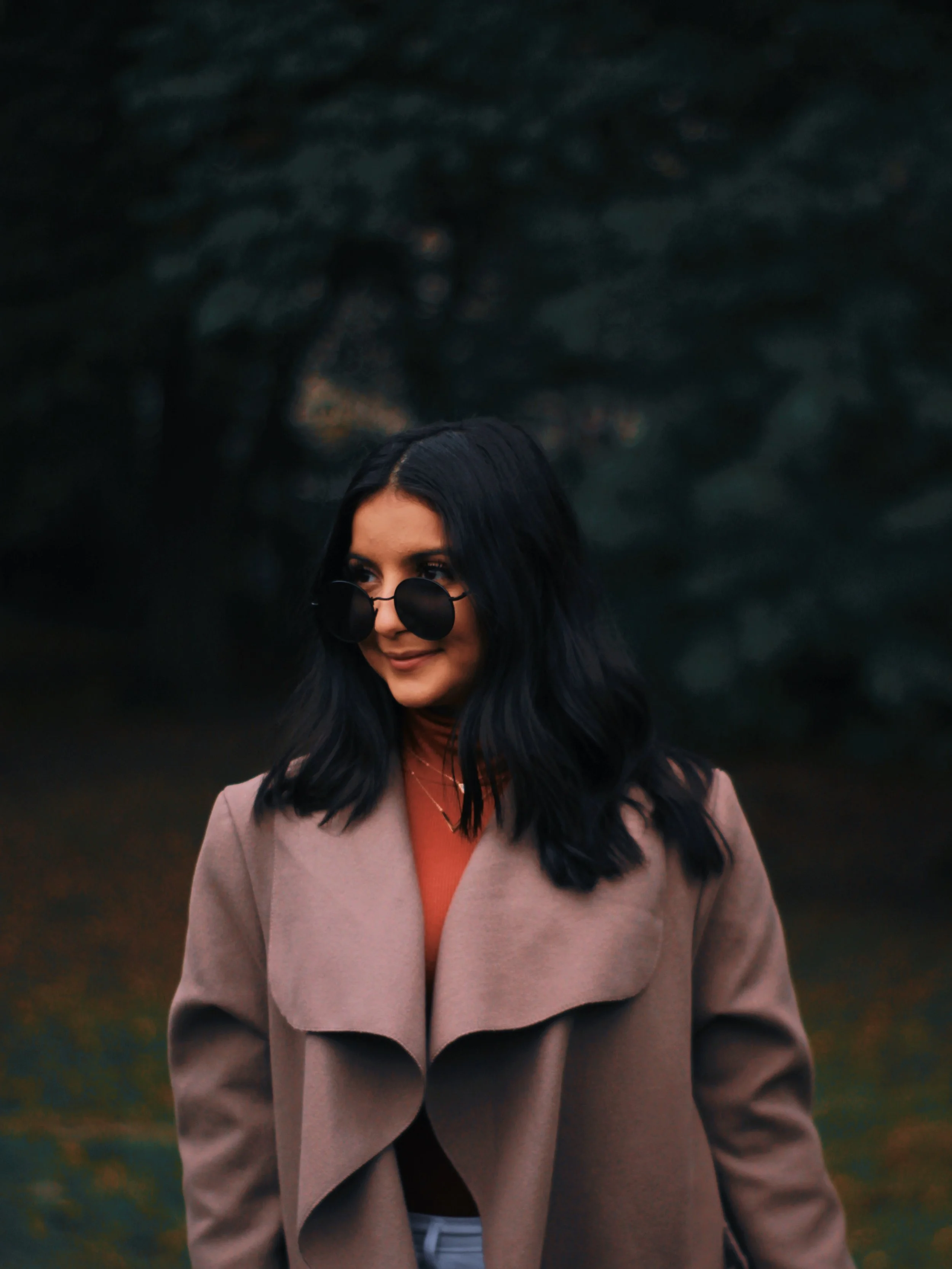 A woman with dark hair wearing oversized black sunglasses, a tan coat, and an orange top, standing outdoors against a dark, blurred background of trees.