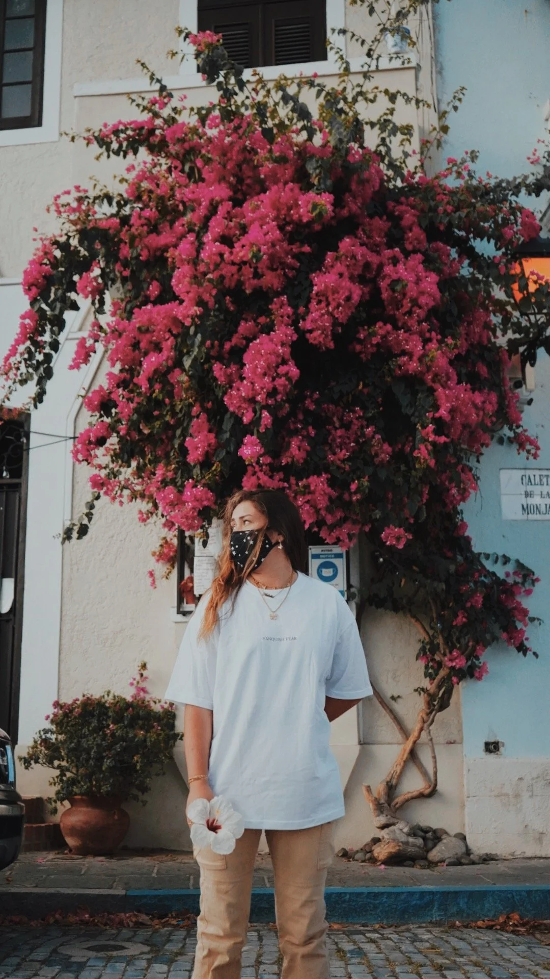 A woman in a white T-shirt, beige pants, and a face mask stands on a cobblestone street in front of a large pink flowering shrub. She holds a white hibiscus flower in her hand and looks to her left.