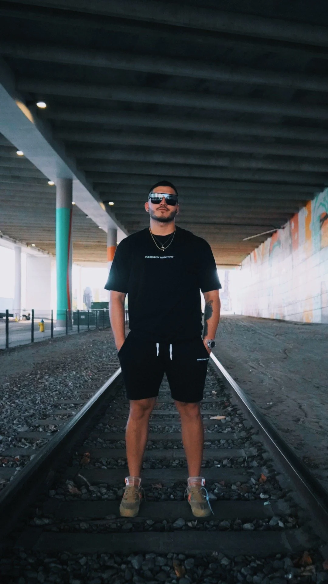 A man standing on train tracks under a bridge, wearing sunglasses, a black T-shirt, black shorts, tan shoes, with jewelry and a watch.