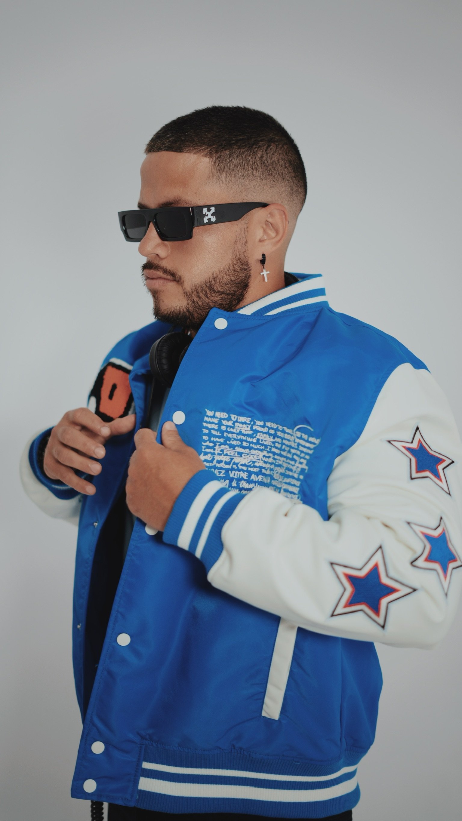 A young man wearing black sunglasses, a blue and white varsity jacket with star patches, and black headphones around his neck.