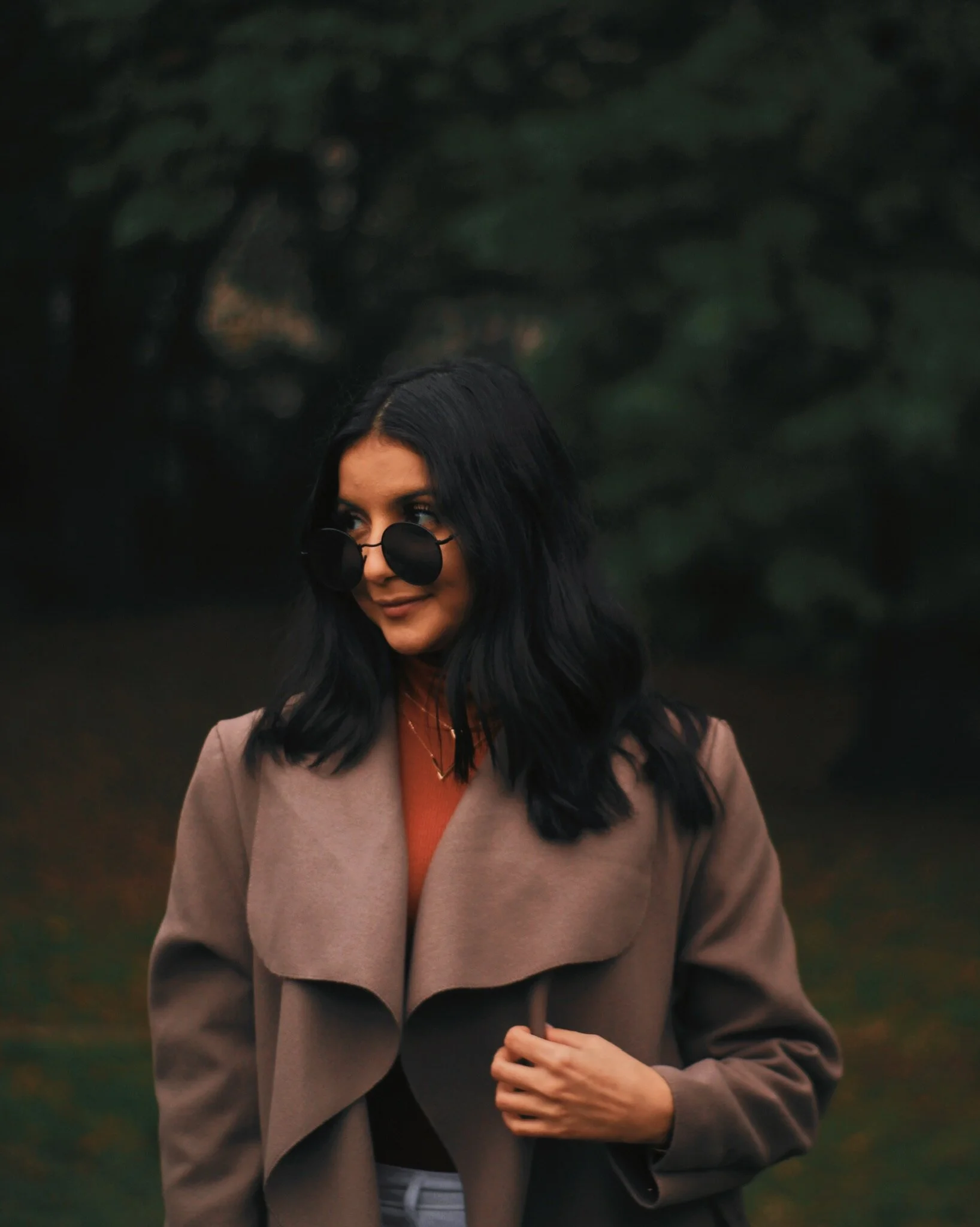 Woman with black hair wearing dark sunglasses, a tan coat, and a rust-colored turtleneck, standing outdoors in front of a blurred green background.