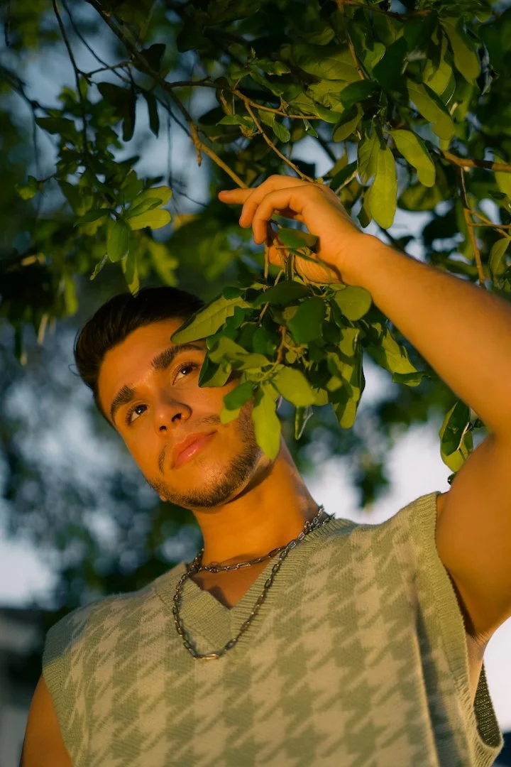 Young man with dark hair and a chain necklace looking at leaves of a tree, standing outdoors during the evening.