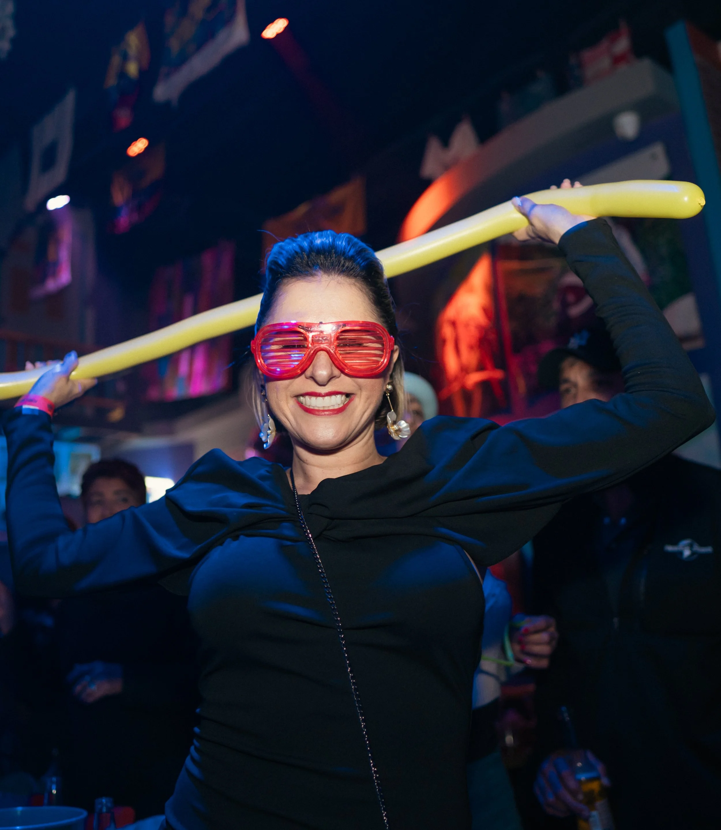 A woman in a black shirt is wearing red shutter shades, smiling, and holding a yellow pool noodle over her shoulders at a lively indoor party or club.