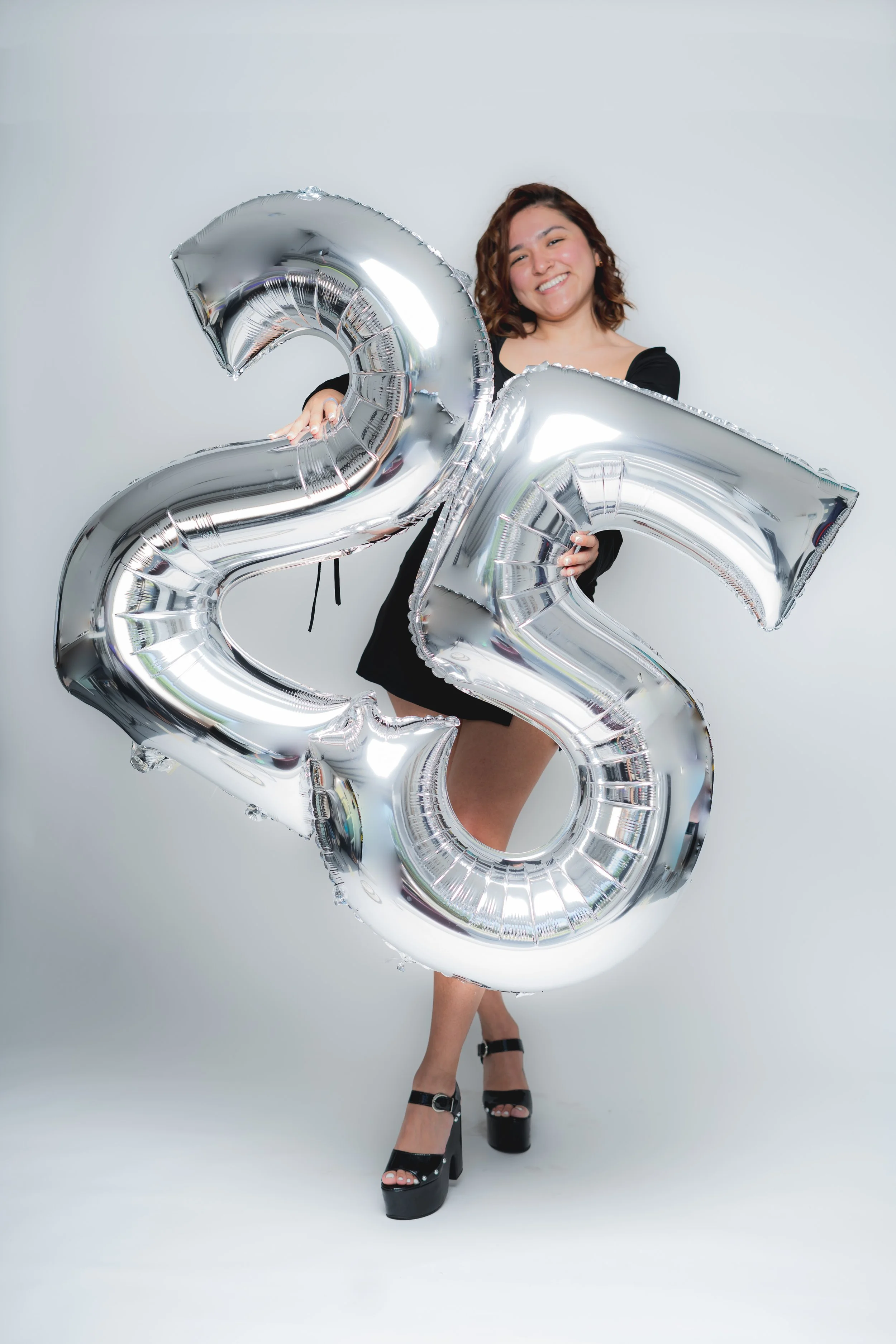 A woman in black shoes and a black dress is smiling and holding large silver balloons shaped as the numbers 2 and 5, celebrating a 25th birthday or anniversary.