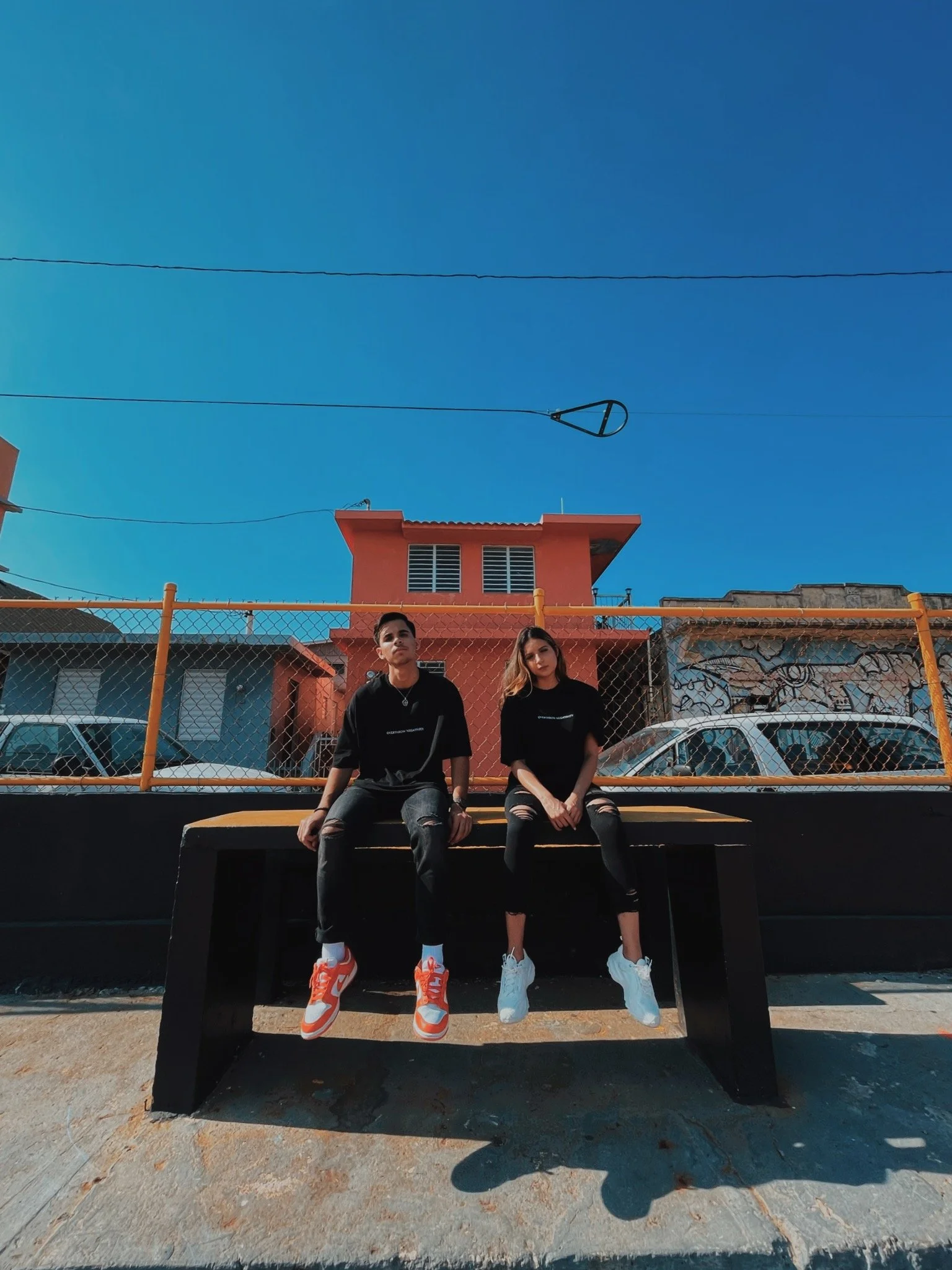 Two young adults, a man and a woman, sitting on a black bench in front of a chain-link fence with a colorful building behind them, under a clear blue sky.