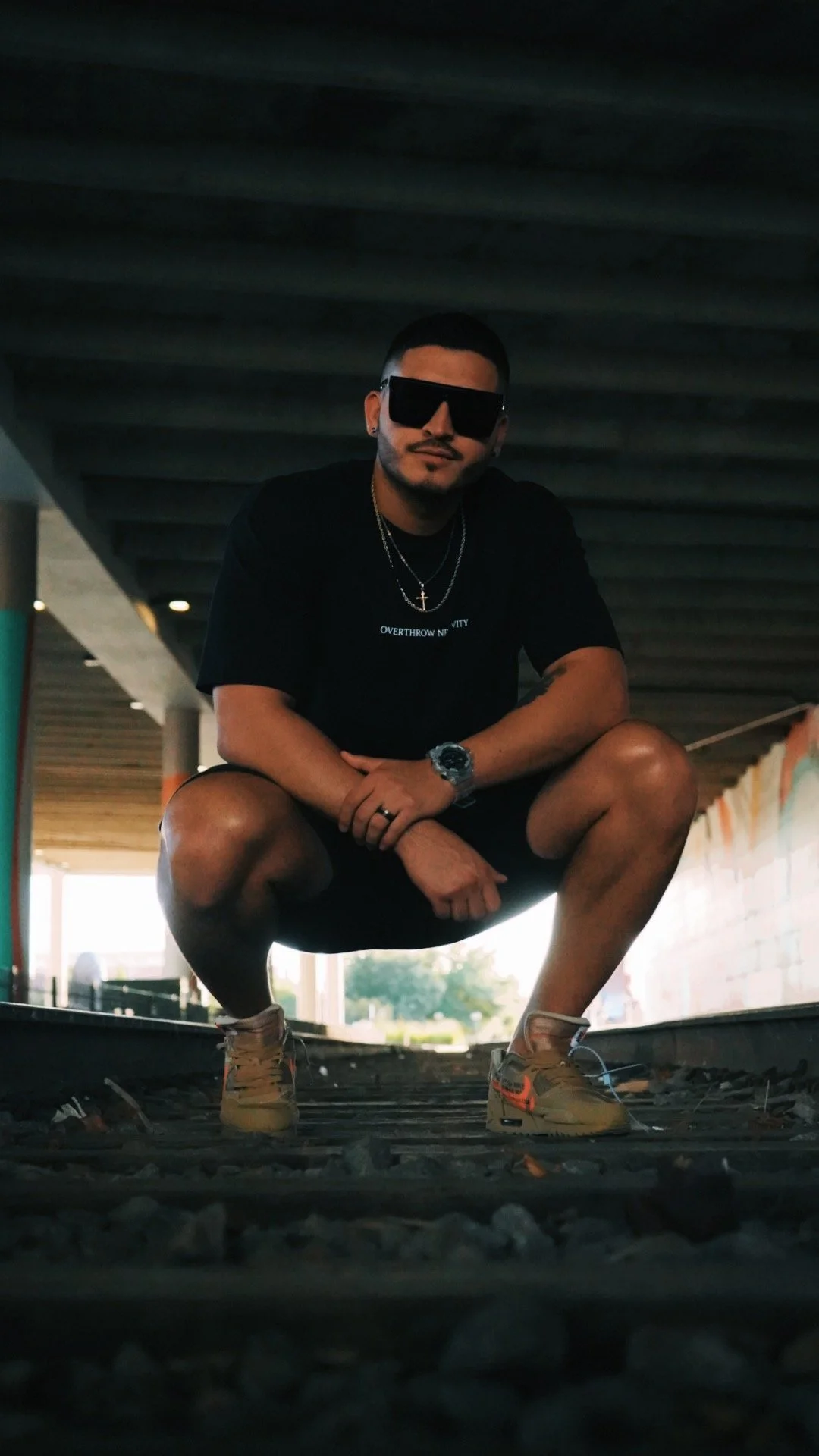 A man squatting on train tracks under a bridge, wearing sunglasses, a black T-shirt, shorts, sneakers, a watch, and necklaces.