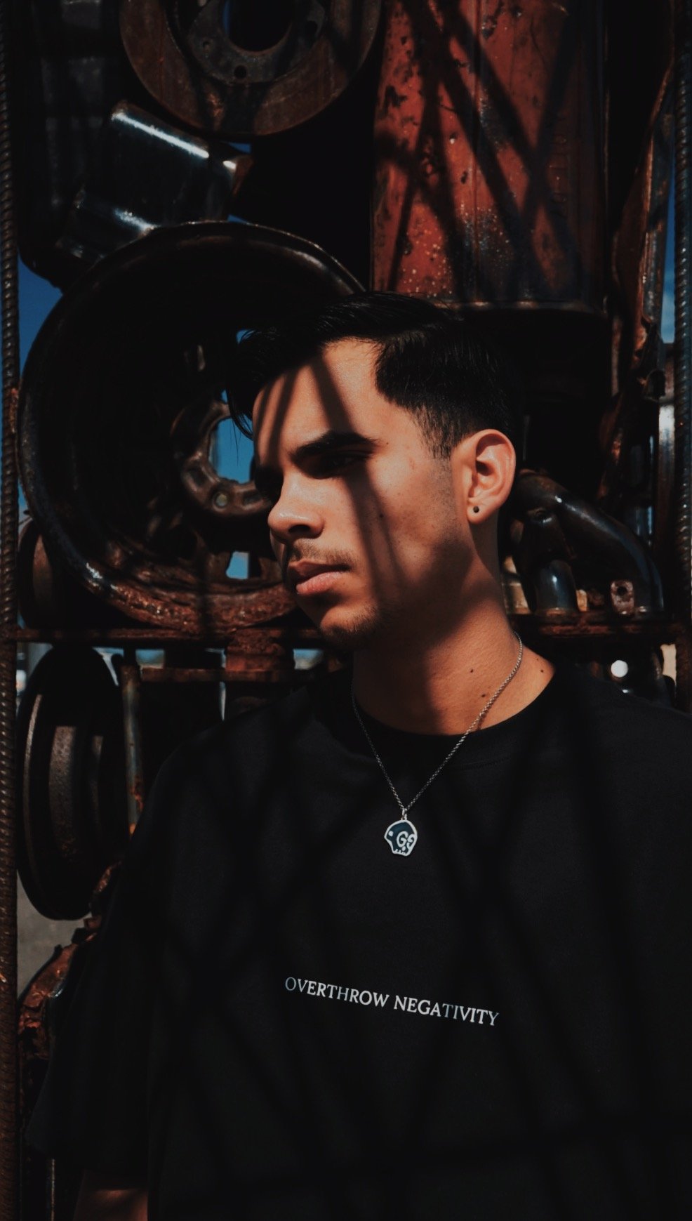 A young man with short dark hair and earrings stands in front of a stack of rusty metal parts, with shadows cast across his face by the metal. He wears a black T-shirt with white text that reads 'OVERTHROW NEGATIVITY' and a necklace with a skull pend