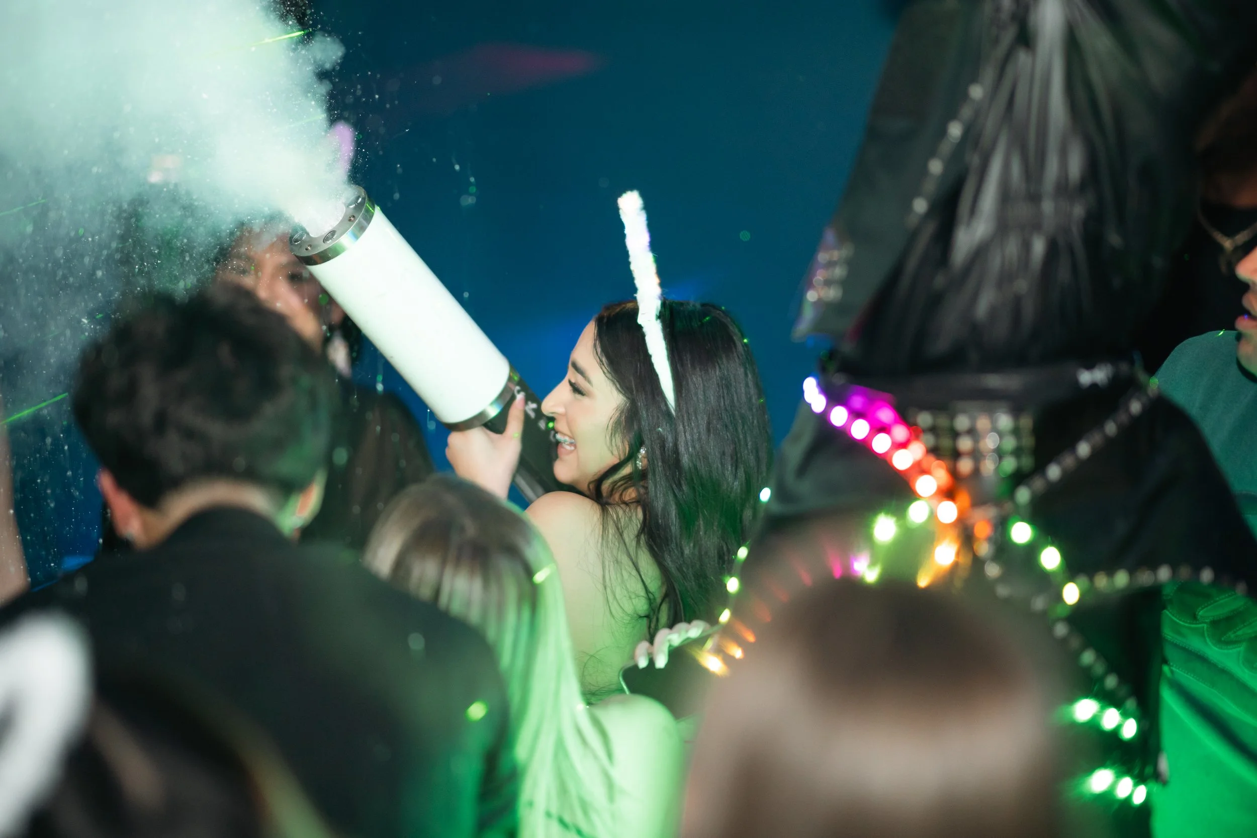 A woman with a unicorn headband smiling and using a party popper on a man at a celebration surrounded by people wearing colorful accessories and lights.
