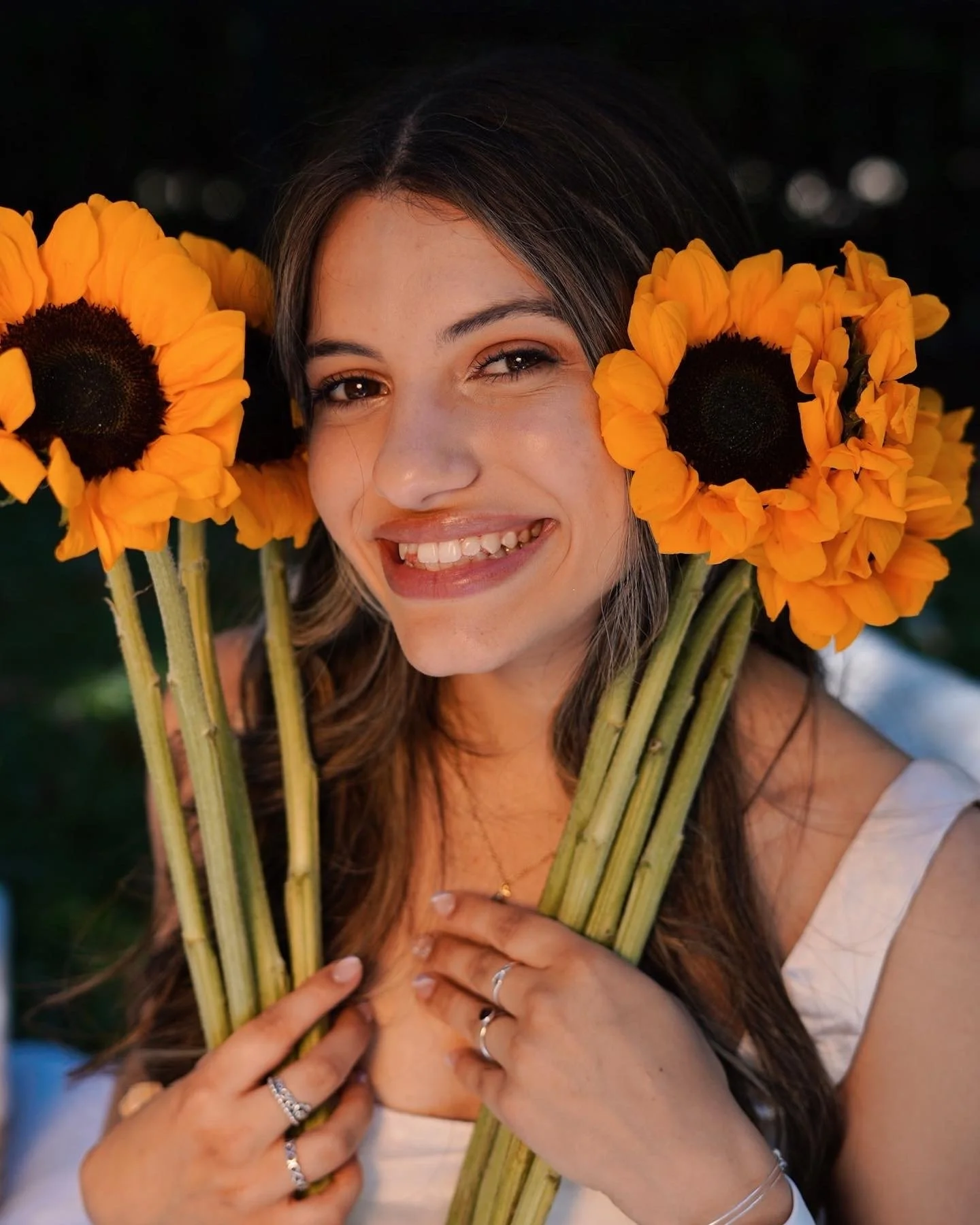 A young woman smiling with sunflowers in her hands, wearing a white top and jewelry.