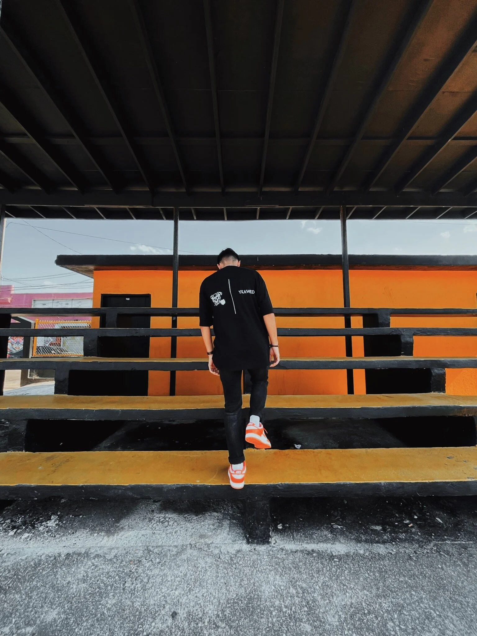 Person wearing black clothing, orange and white sneakers ascending yellow and black stairs, with an orange wall behind him and a dark overhang above.