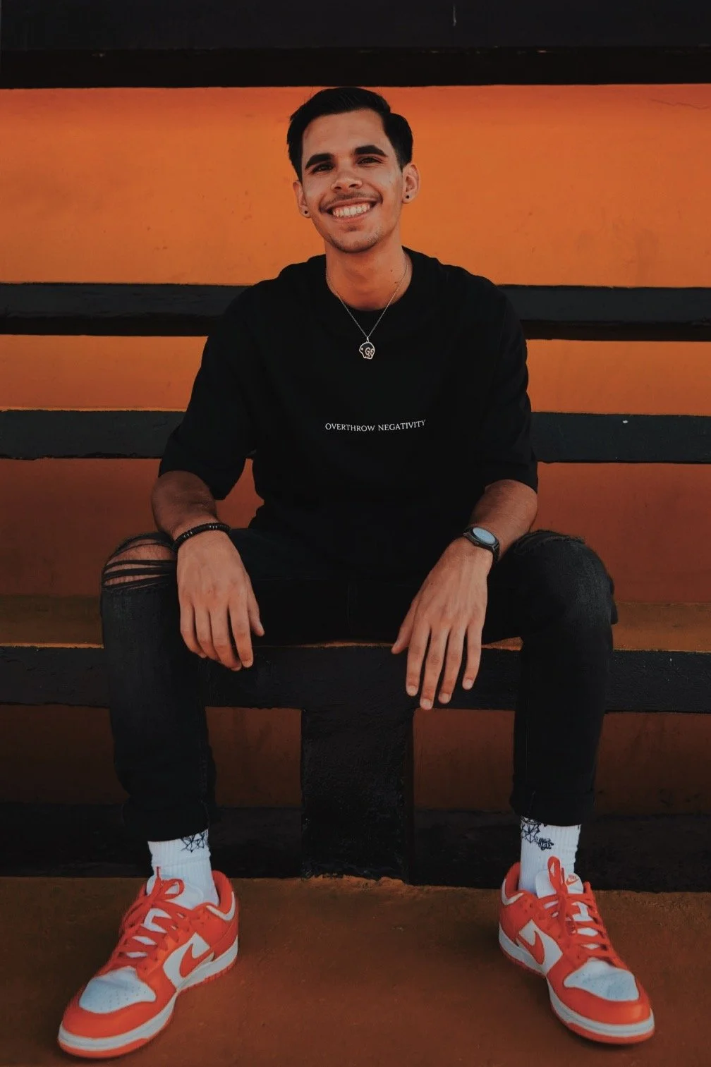A young man with dark hair, wearing a black shirt with white text, ripped black jeans, bright orange and white sneakers, white socks, and accessories, sitting on a black bench against an orange and black wall, smiling at the camera.