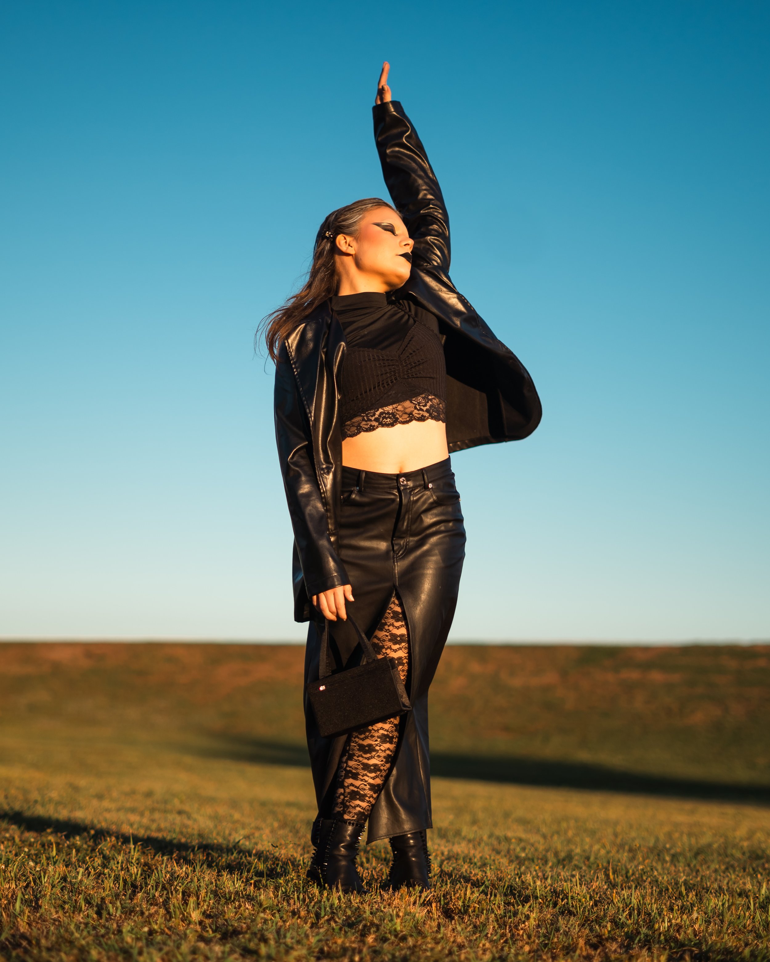A woman in black leather outfit, including a jacket, skirt, and lace tights, standing outdoors on grass during sunset, with her right arm raised and her eyes closed.