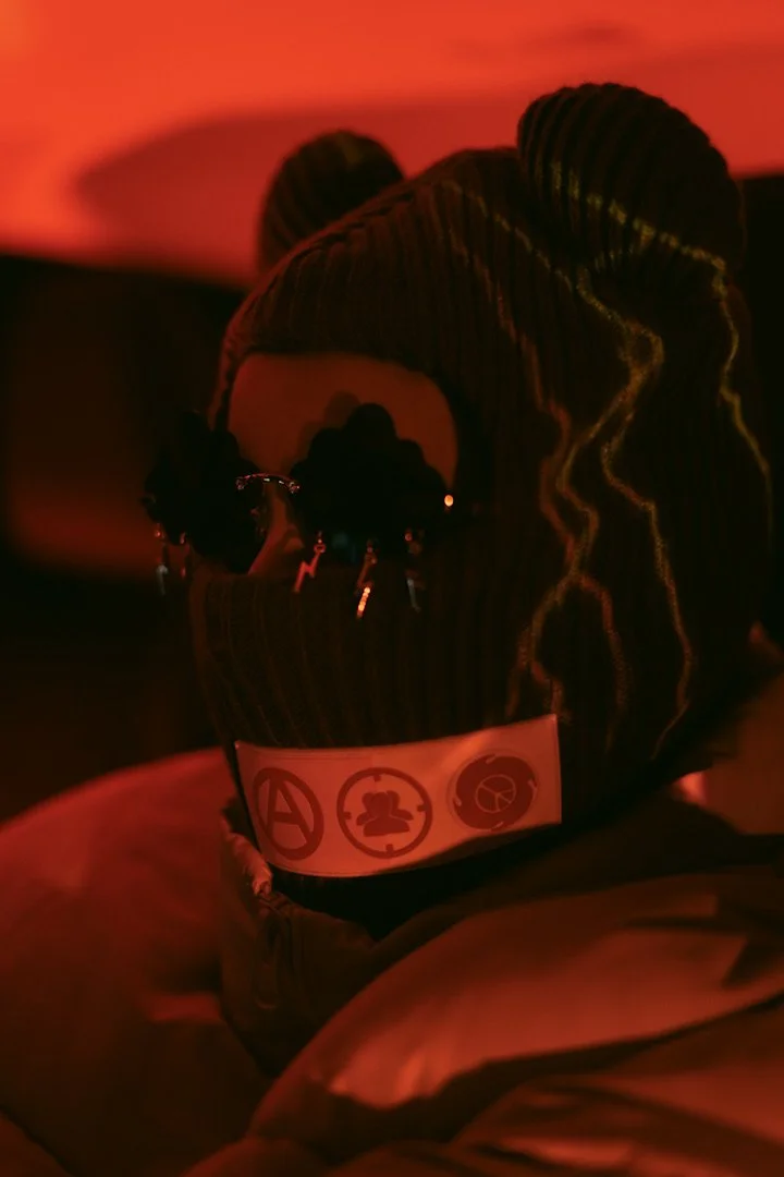 Person wearing a beanie and sunglasses, with a strap featuring social and political icons, in a dimly lit setting with red/orange lighting.