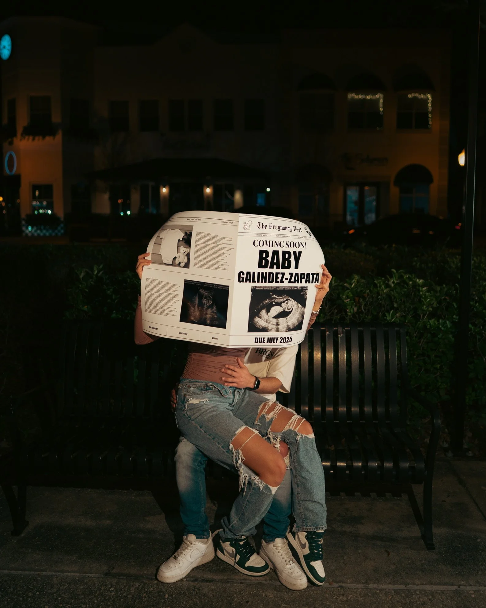 Two people sitting on a bench at night, holding a newspaper with a prenatal ultrasound image and a baby announcement, with one person's hand on lap and the other person's arm visible.