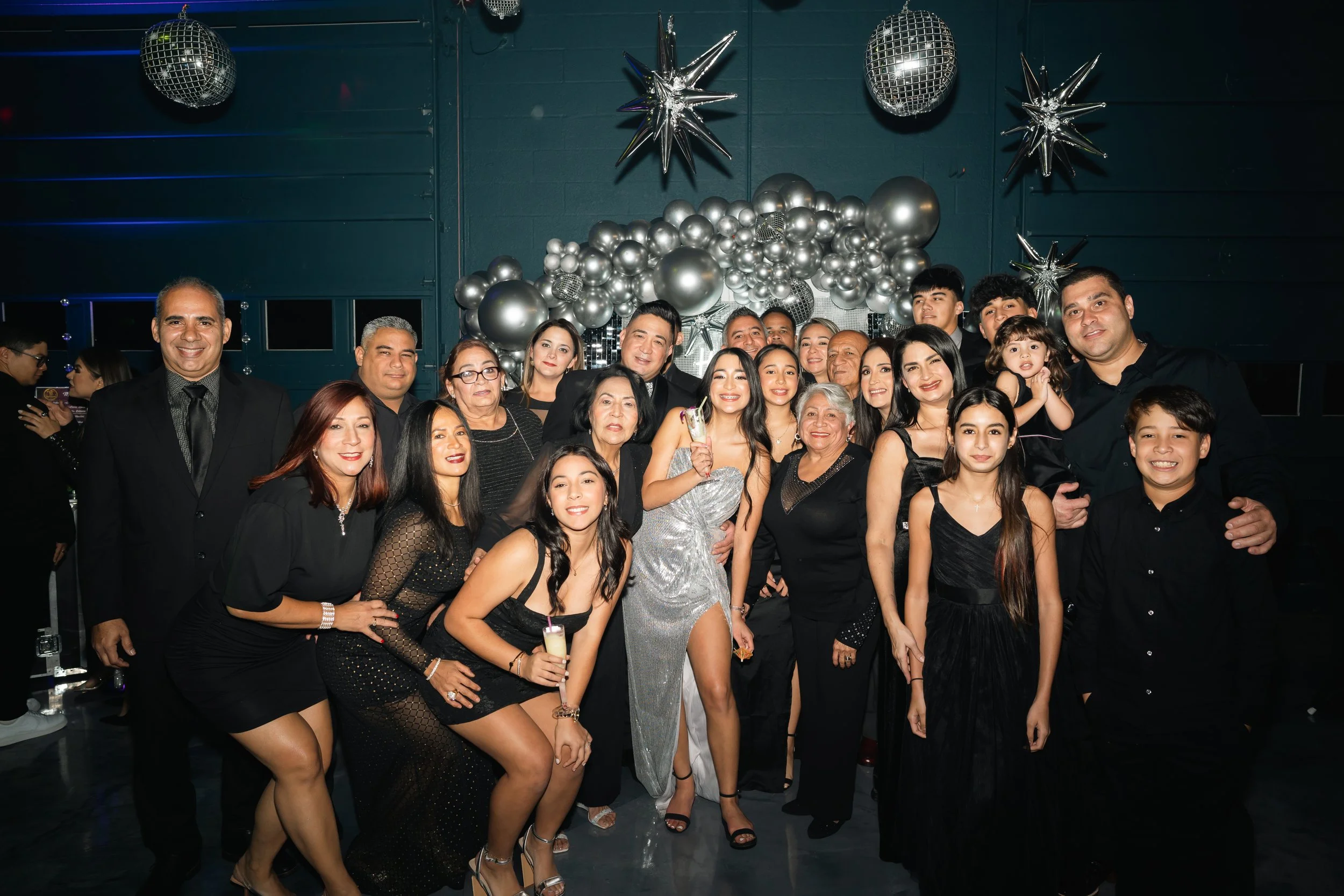 A large group of people celebrating at a party in front of a silver and black balloon backdrop with star and disco ball decorations. Everyone is dressed in black or silver, smiling and holding drinks.