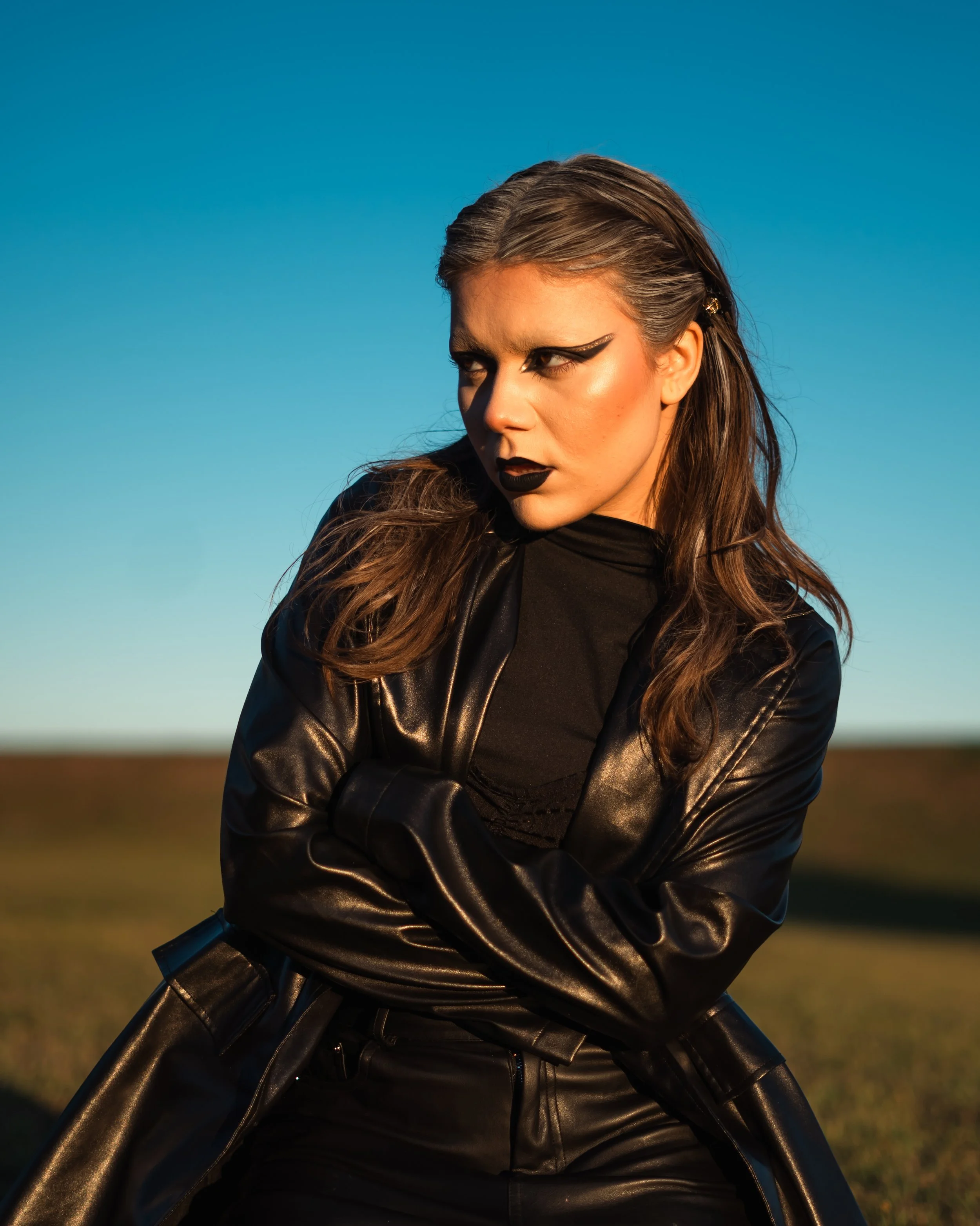 A woman with dark makeup and leather clothing poses outdoors against a clear blue sky.