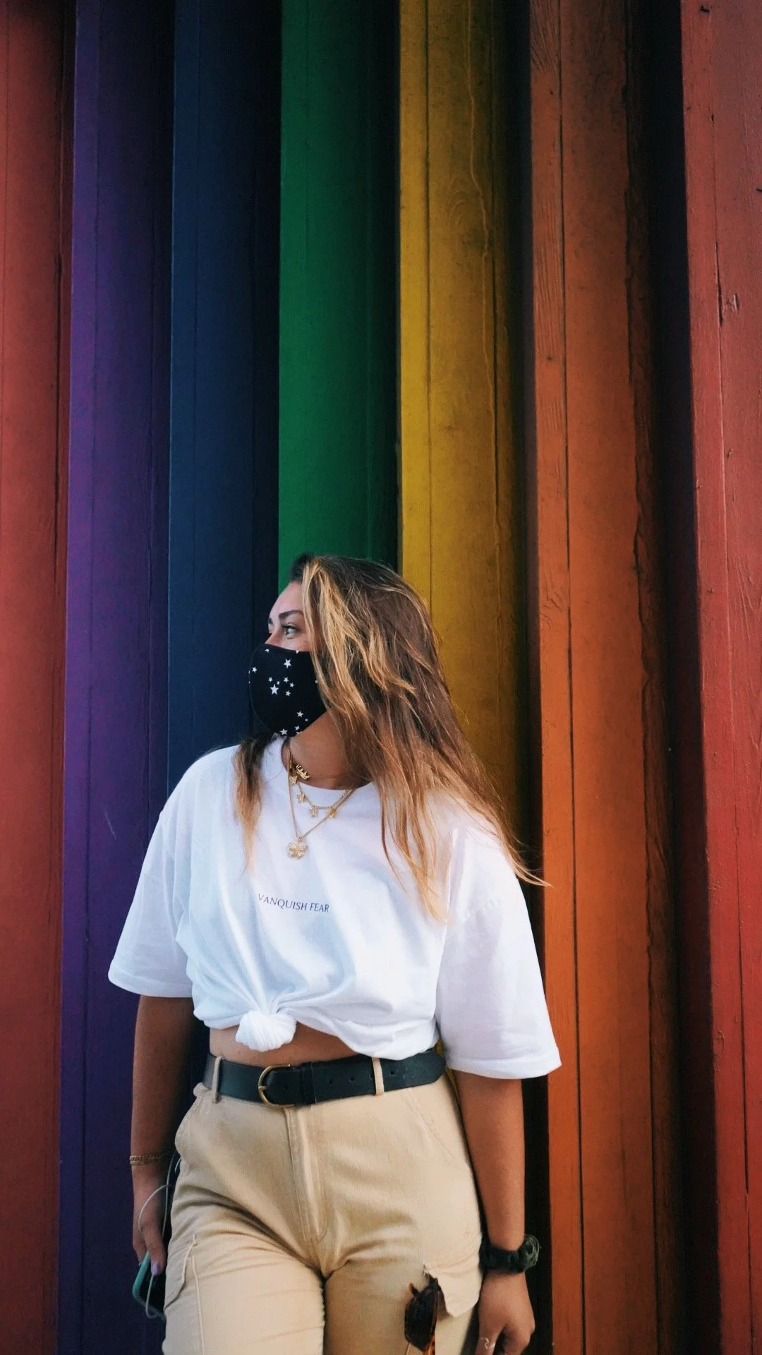 A woman wearing a black face mask with white stars, a white t-shirt tied at the waist, beige high-waisted pants, standing in front of a rainbow-colored wooden wall.