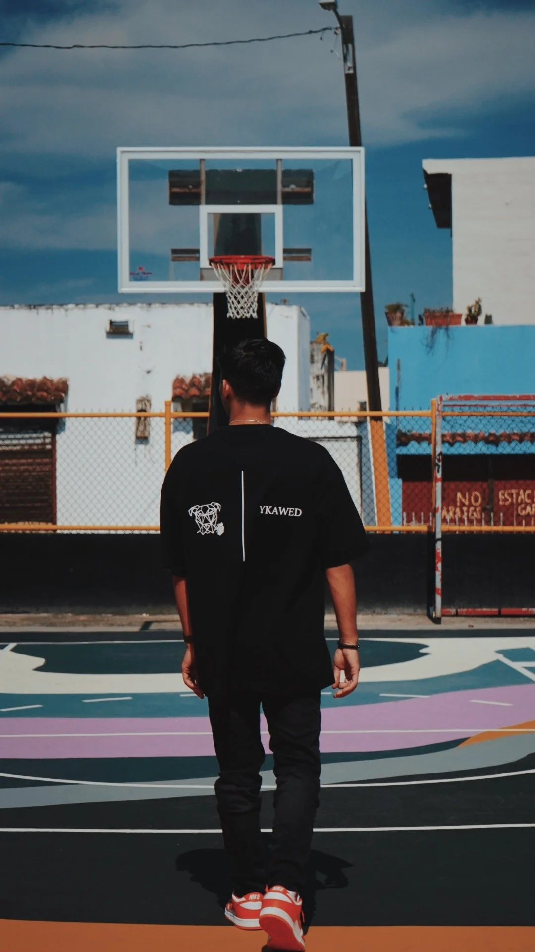 A young man in a black T-shirt with white logos on the back walks towards a basketball hoop on an outdoor court, with colorful markings on the ground and a yellow fence in the background.