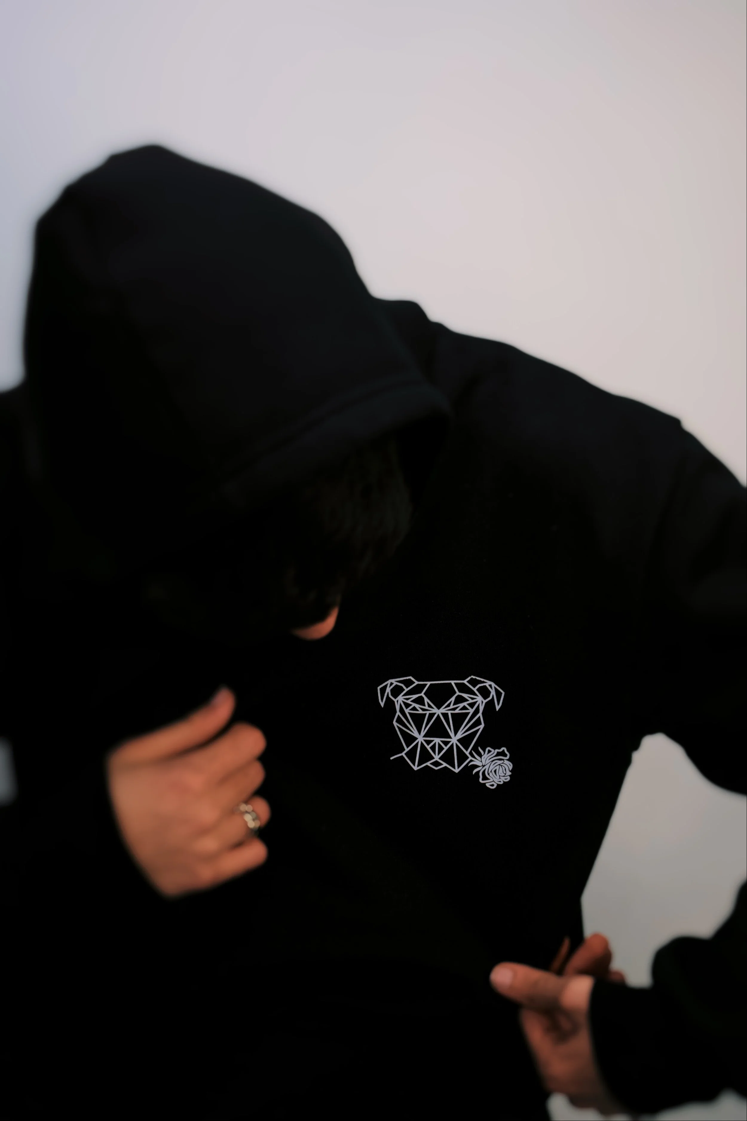 Person wearing a black hoodie with a geometric dog face and rose design on the chest, touching their jacket.