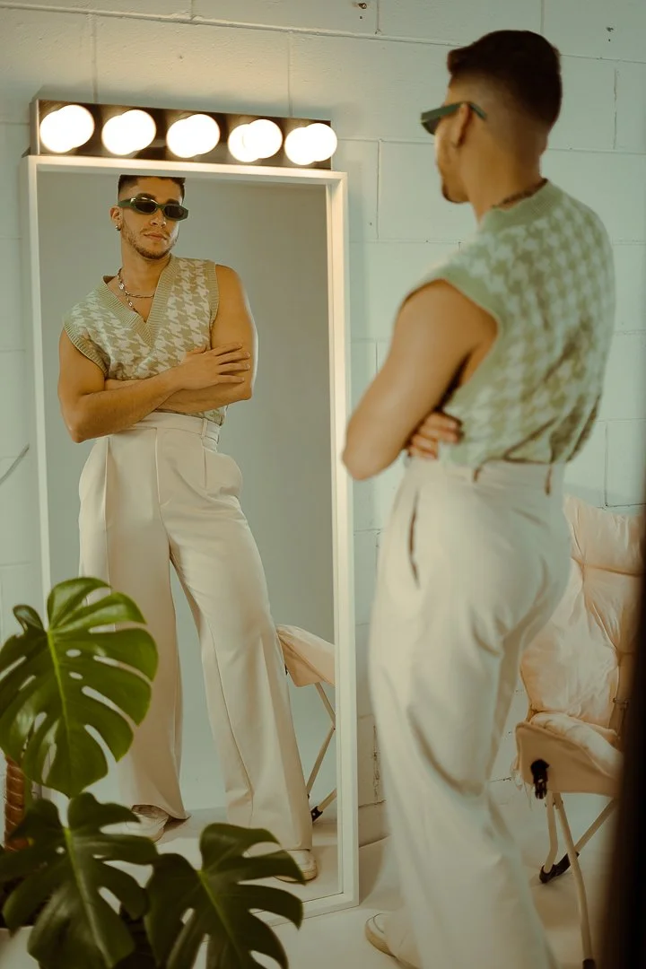 A man with sunglasses looking at his reflection in a mirror, standing with arms crossed. The mirror is in a room with white brick walls and a row of bright lights above. There is green foliage in the foreground and a pink cushioned chair in the backg