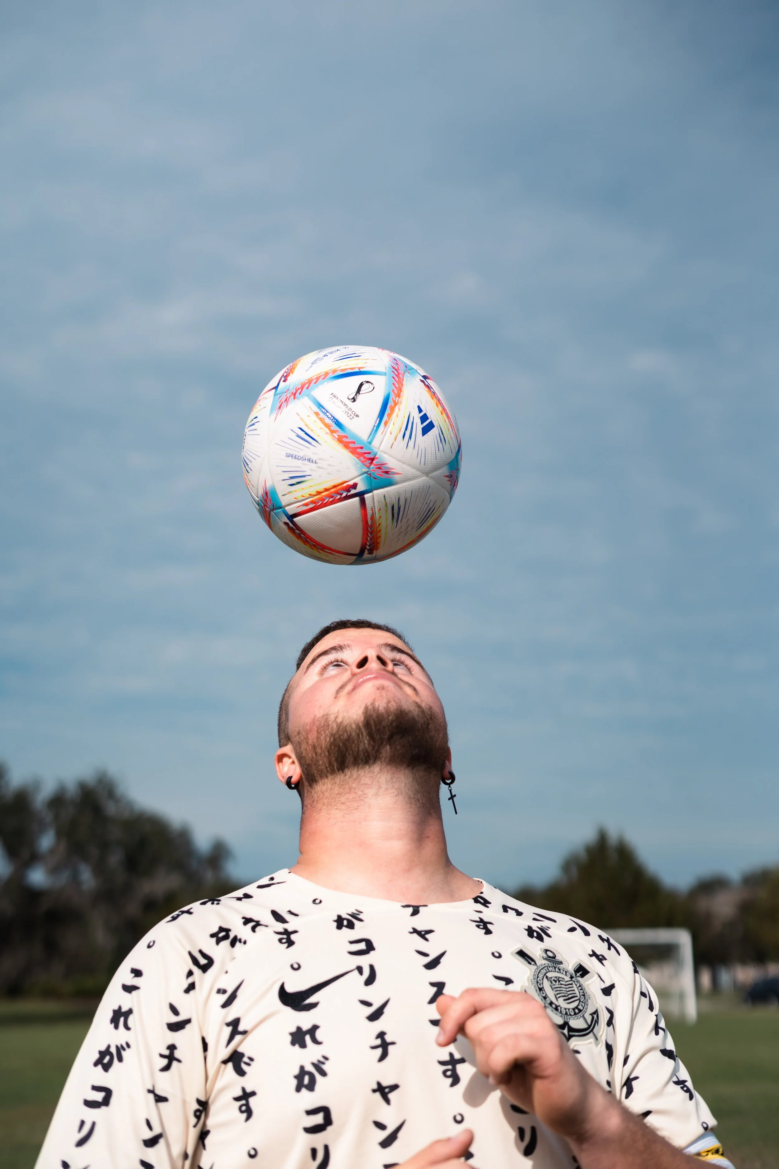 A man with a beard and earrings is playing soccer outdoors during daytime, heading a colorful soccer ball against a cloudy sky background.