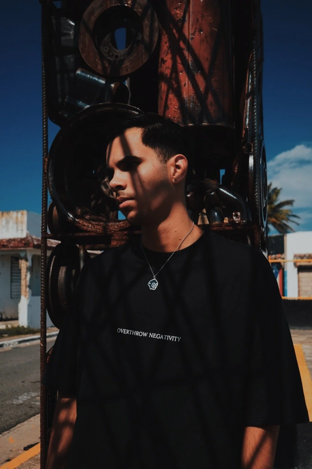 A young man with short dark hair and earrings, wearing a black t-shirt with the words "OVERTHROW NEGATIVITY" on it, standing outdoors against rusted metal equipment, with a partly cloudy sky and some buildings in the background.