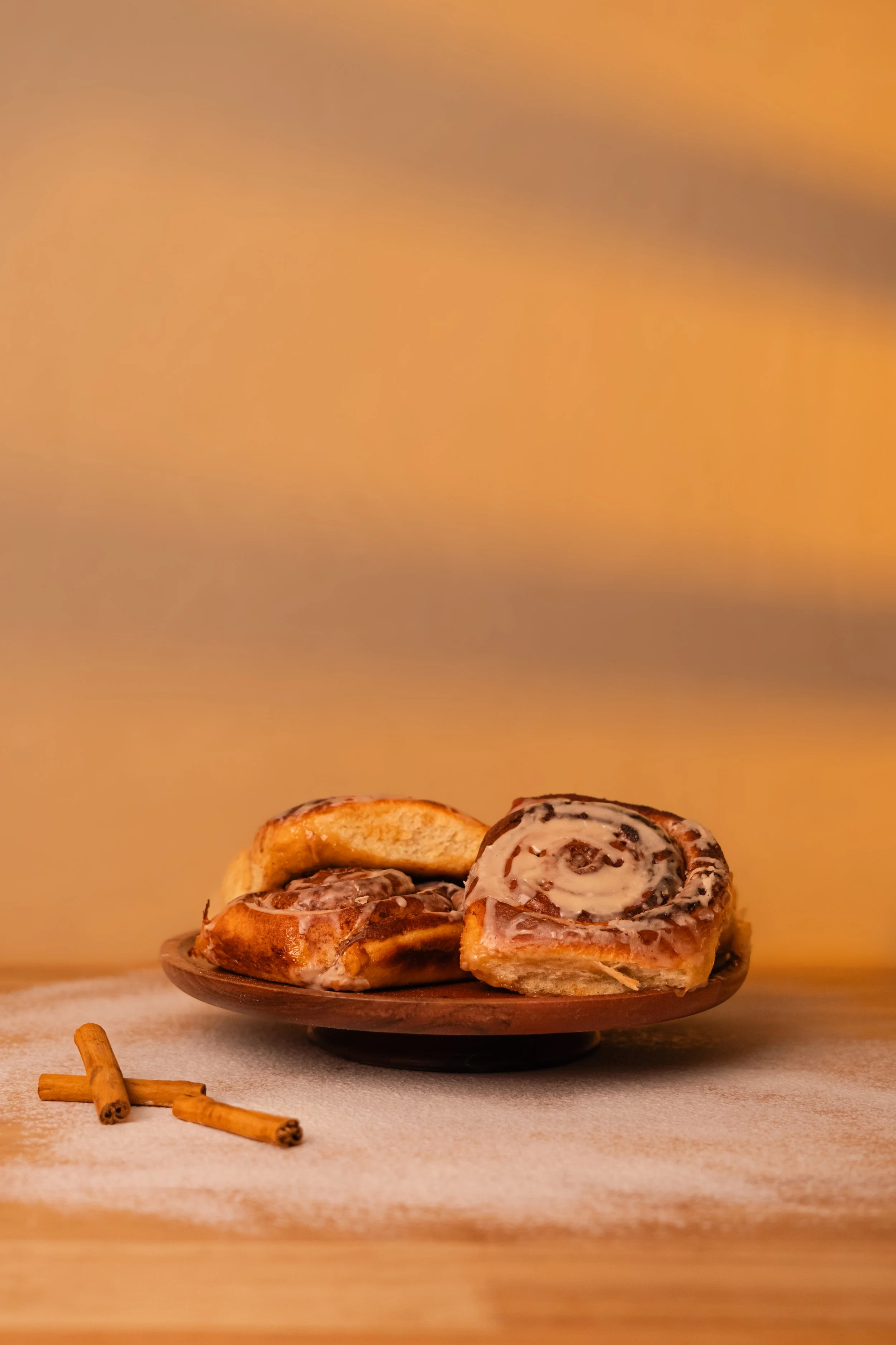 Cinnamon rolls with icing on a dark plate, with cinnamon sticks on a light surface and a plain beige background.